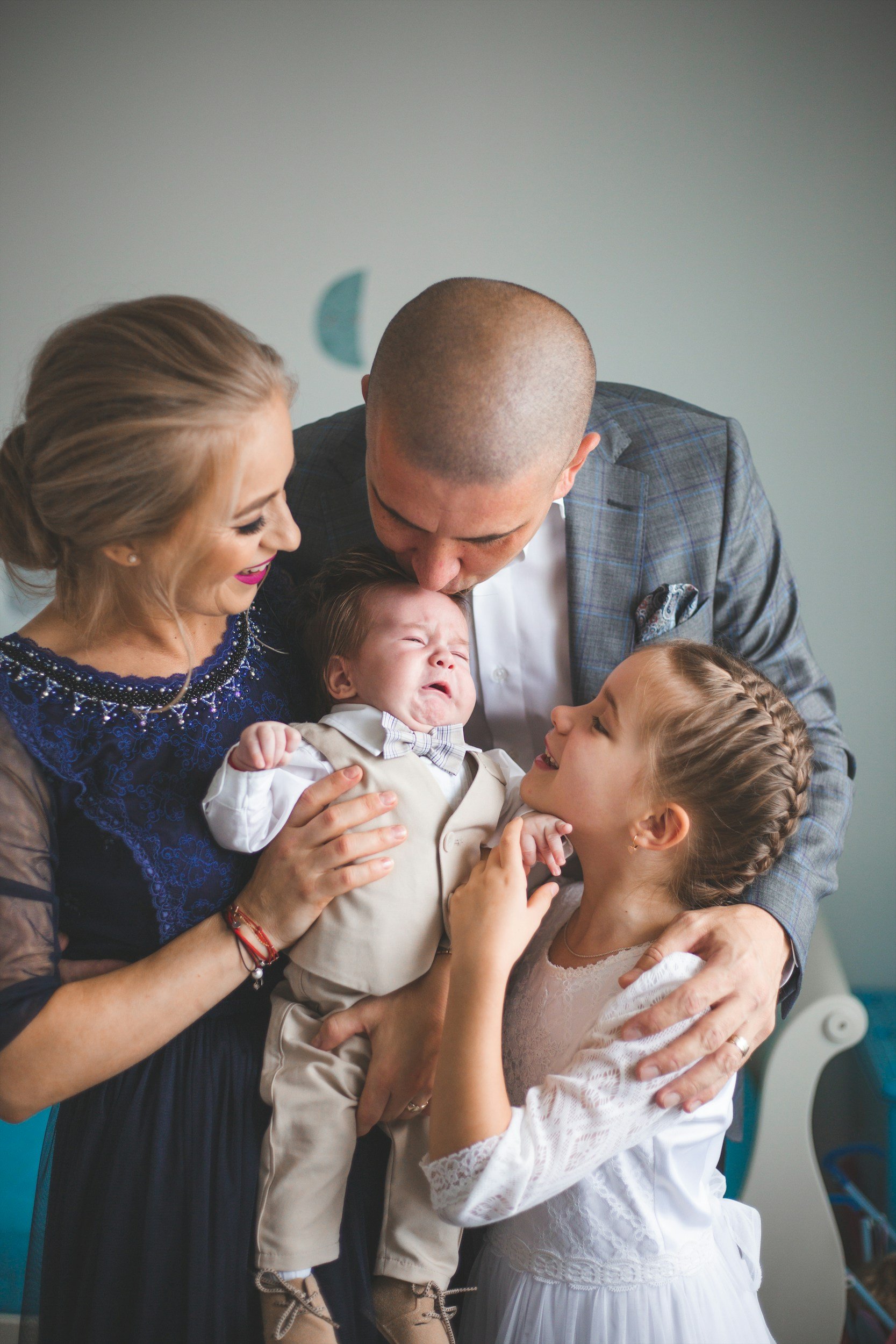 Family with two parents and two children, including a crying baby, in a warm embrace indoors.