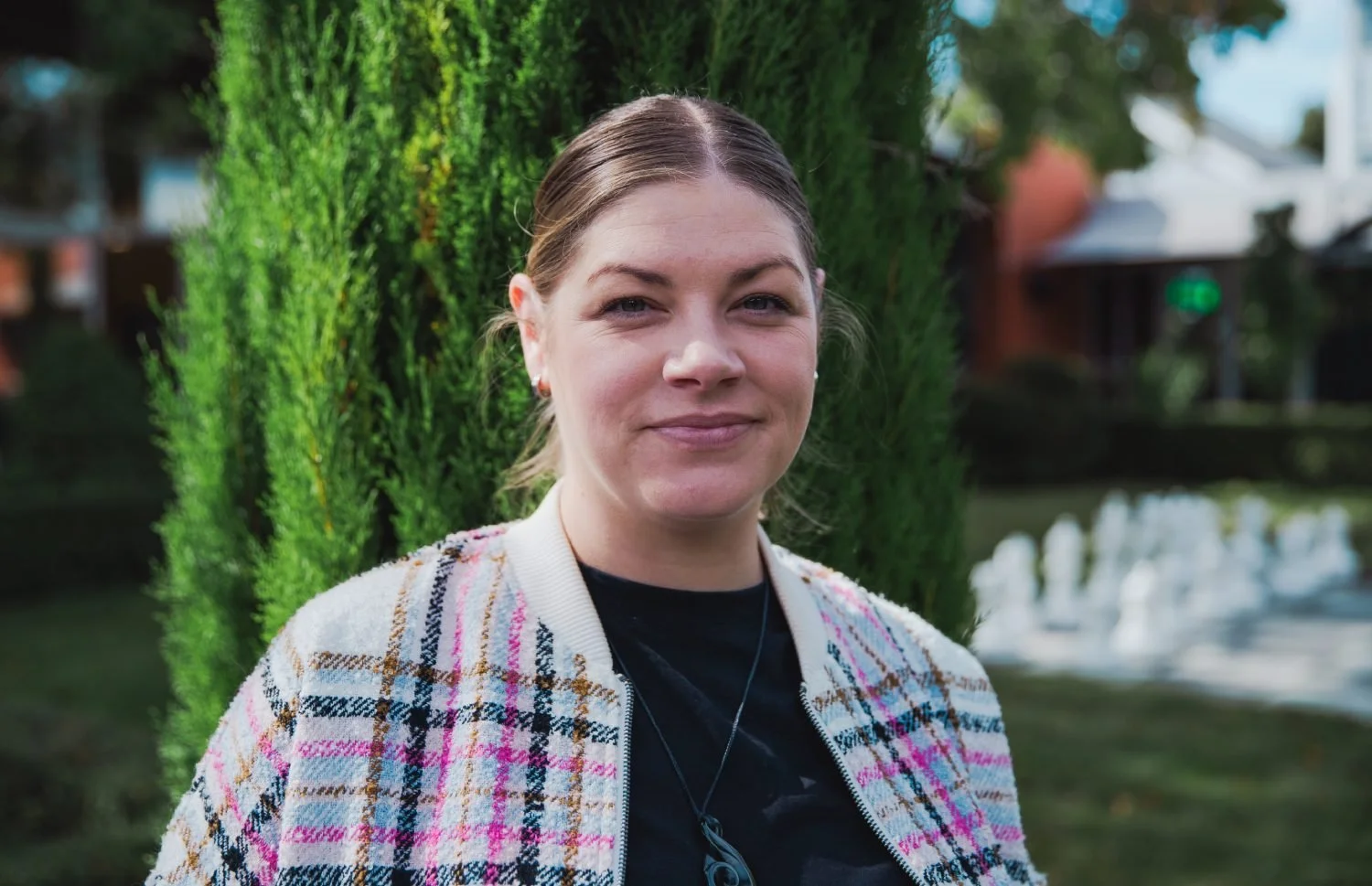 Woman standing outdoors with green foliage background, wearing a plaid jacket and a black shirt.