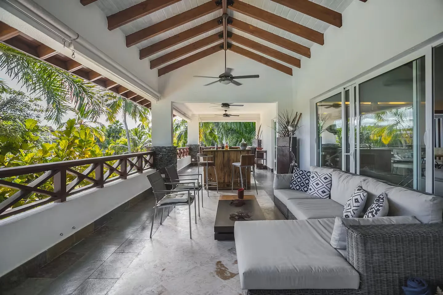 Amplia terraza con muebles de exterior, incluyendo sillones de mimbre, una mesa de centro, una barra de bar en el fondo, y una vista a un entorno con muchas plantas y árboles verdes.
