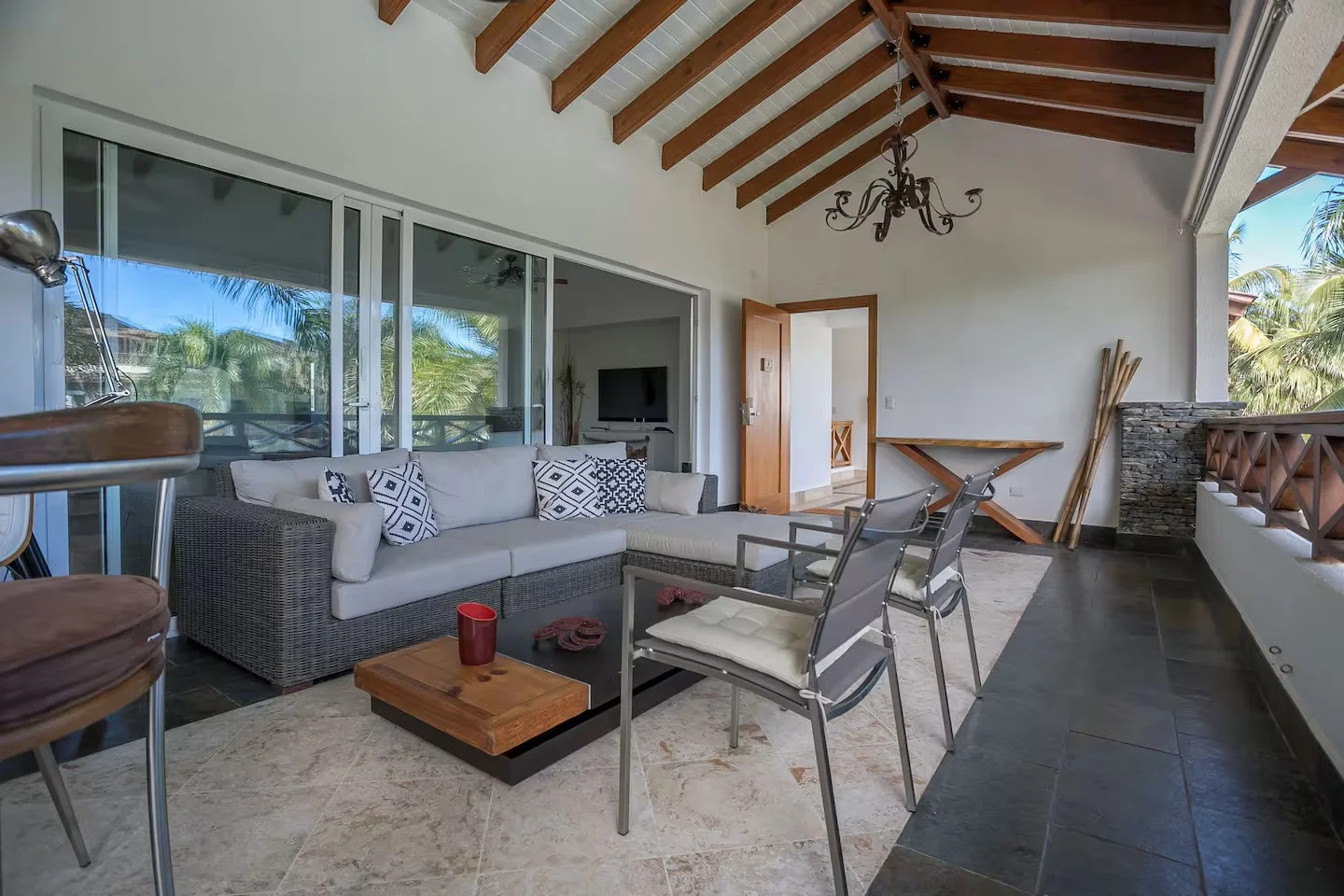 Sala de estar con sofá gris, sillas metálicas, mesa de centro de madera, televisión, paredes blancas, techo de madera, y un balcón con vista a palmeras.