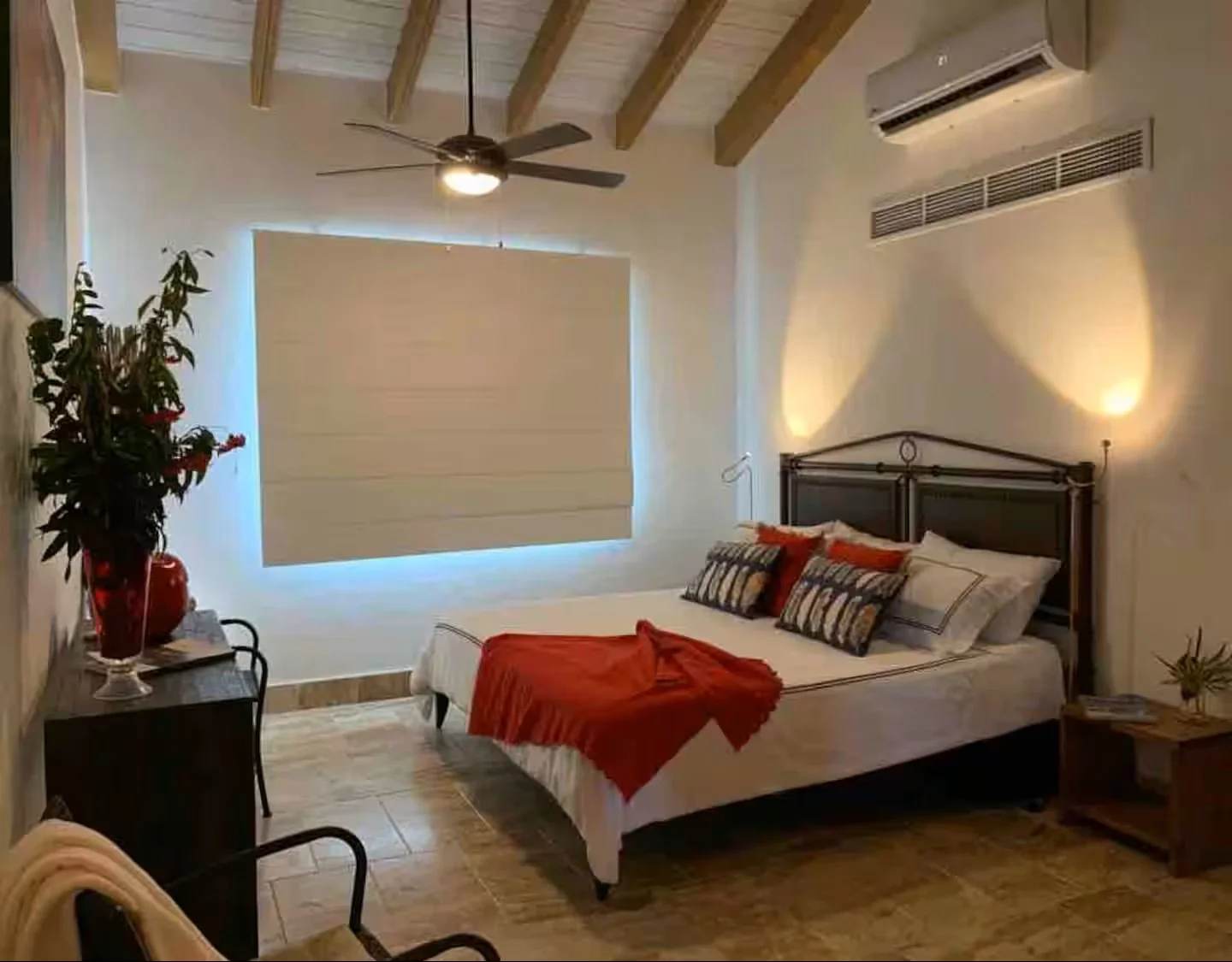 Habitación con cama, almohadas decorativas, ventilador en el techo, aire acondicionado, cortinas semitransparentes, alfombra y muebles de madera, decorada con plantas y lámparas de pared.