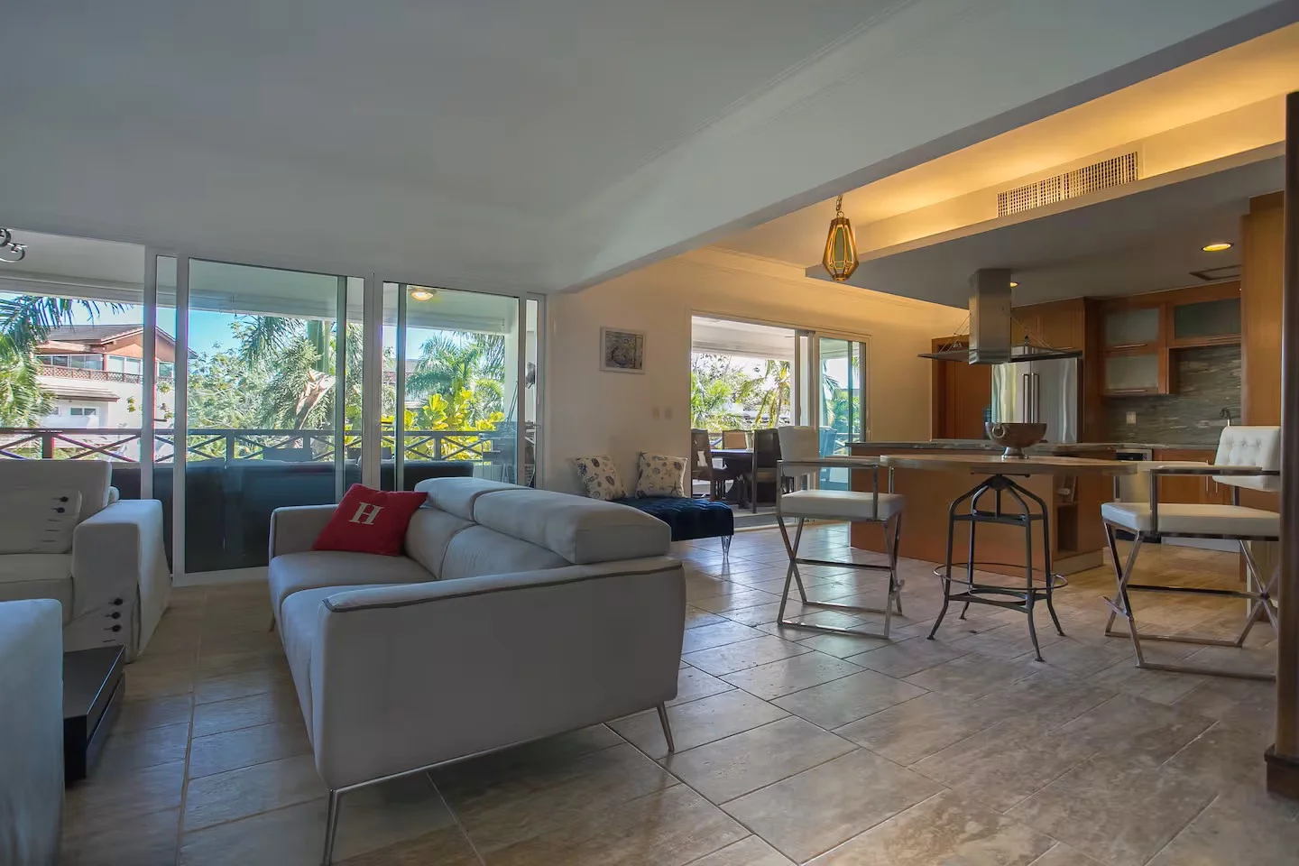 Sala de estar con sillones blancos, cojines decorativos, cocina moderna con barra y sillas, balcón con vista a áreas verdes