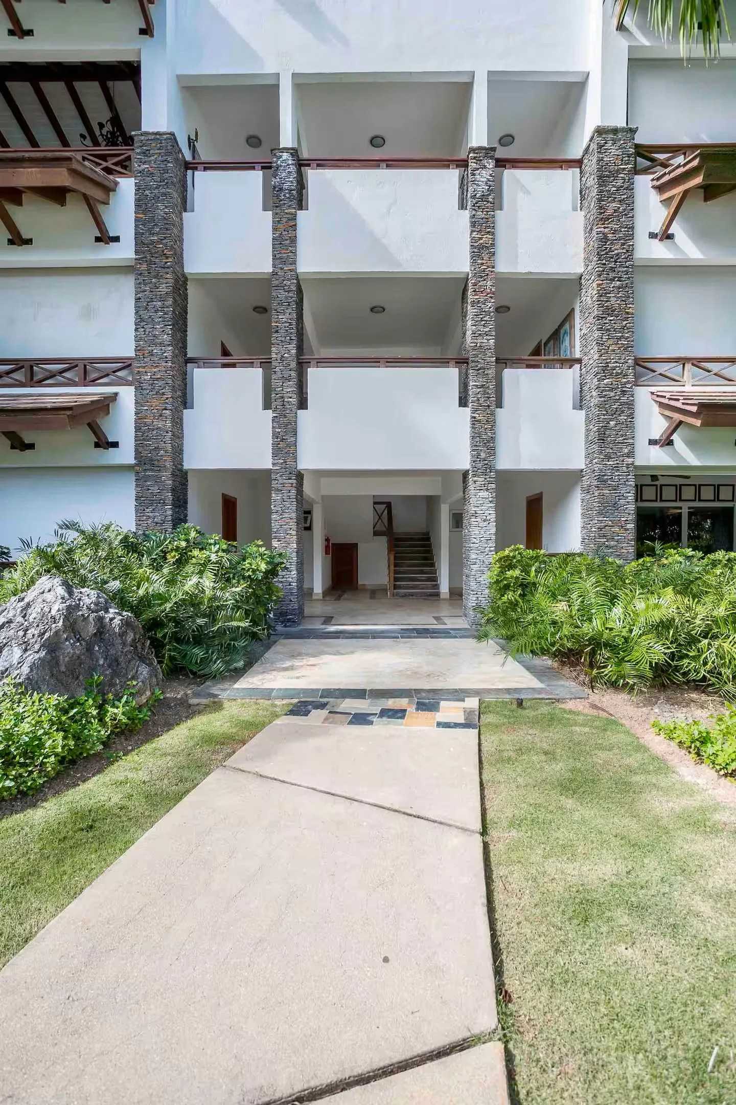 Fachada de un edificio de apartamentos con balcones de madera y columnas de piedra, entrada con sendero de concreto y jardín con plantas verdes.