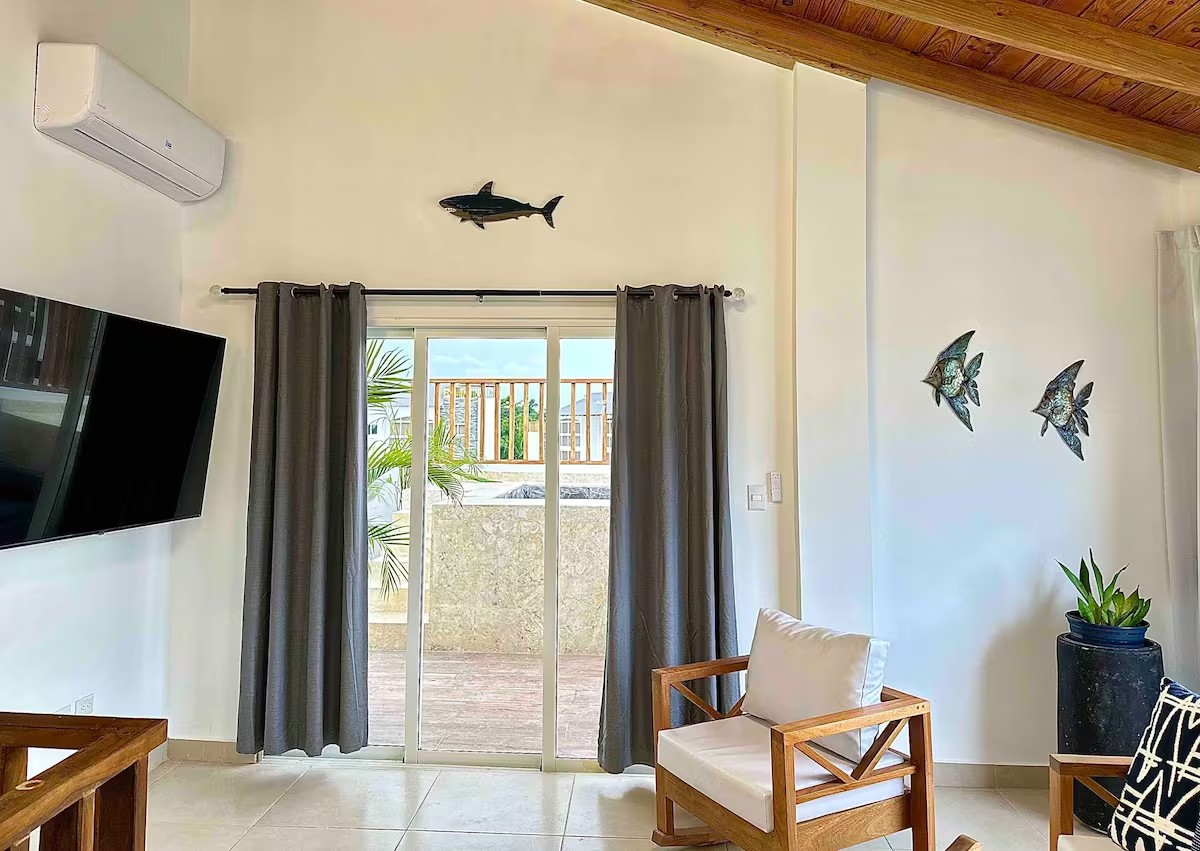 Modern living room with wooden furniture, wall art of fish, air conditioner, TV, sliding glass doors with curtains, and outdoor view.