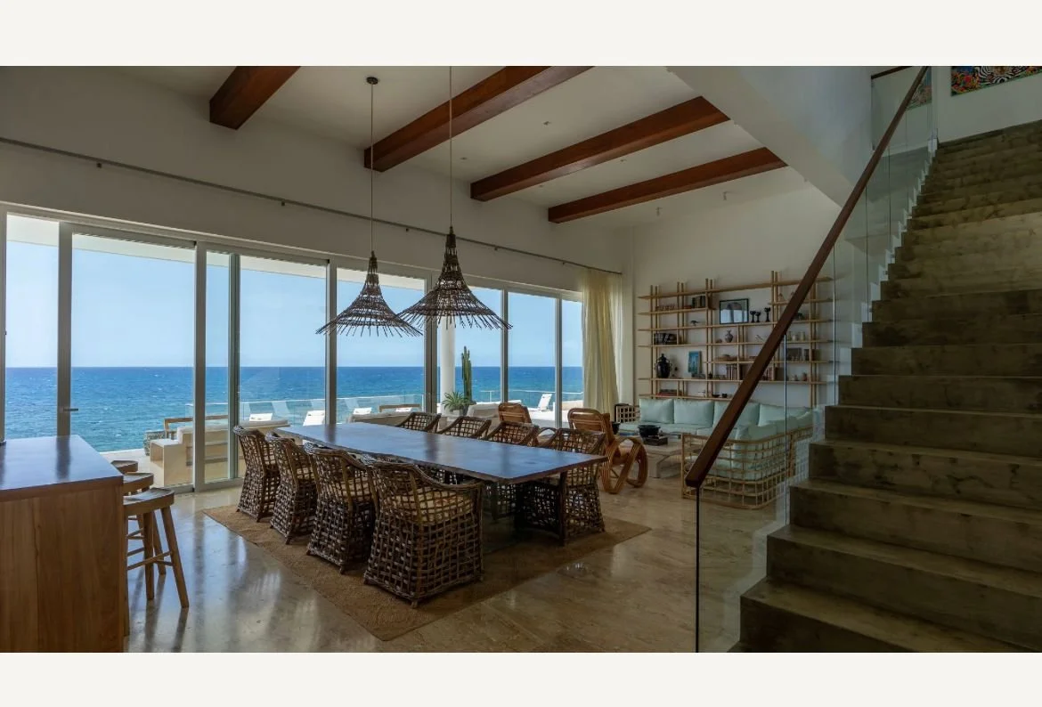 Sala de estar elegante con vista al océano, grandes ventanales, mesa de comedor de madera con sillas de mimbre, lámparas colgantes y escalera moderna, decoración en estilo contemporáneo.