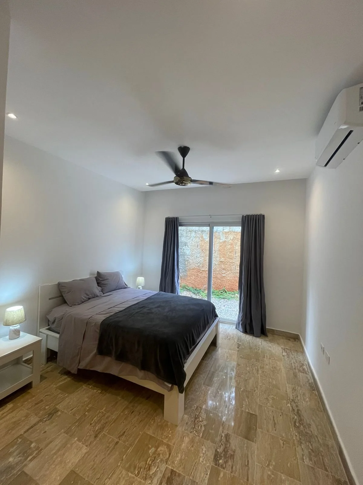 Minimalist bedroom with double bed, ceiling fan, air conditioner, and sliding glass doors with curtains.
