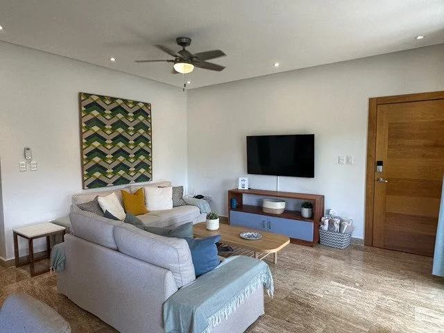 Modern living room with a ceiling fan, L-shaped sofa, and wall-mounted TV. Decor includes geometric wall art, coffee table with plants, and wooden furniture. Natural light and neutral tones create a cozy atmosphere.
