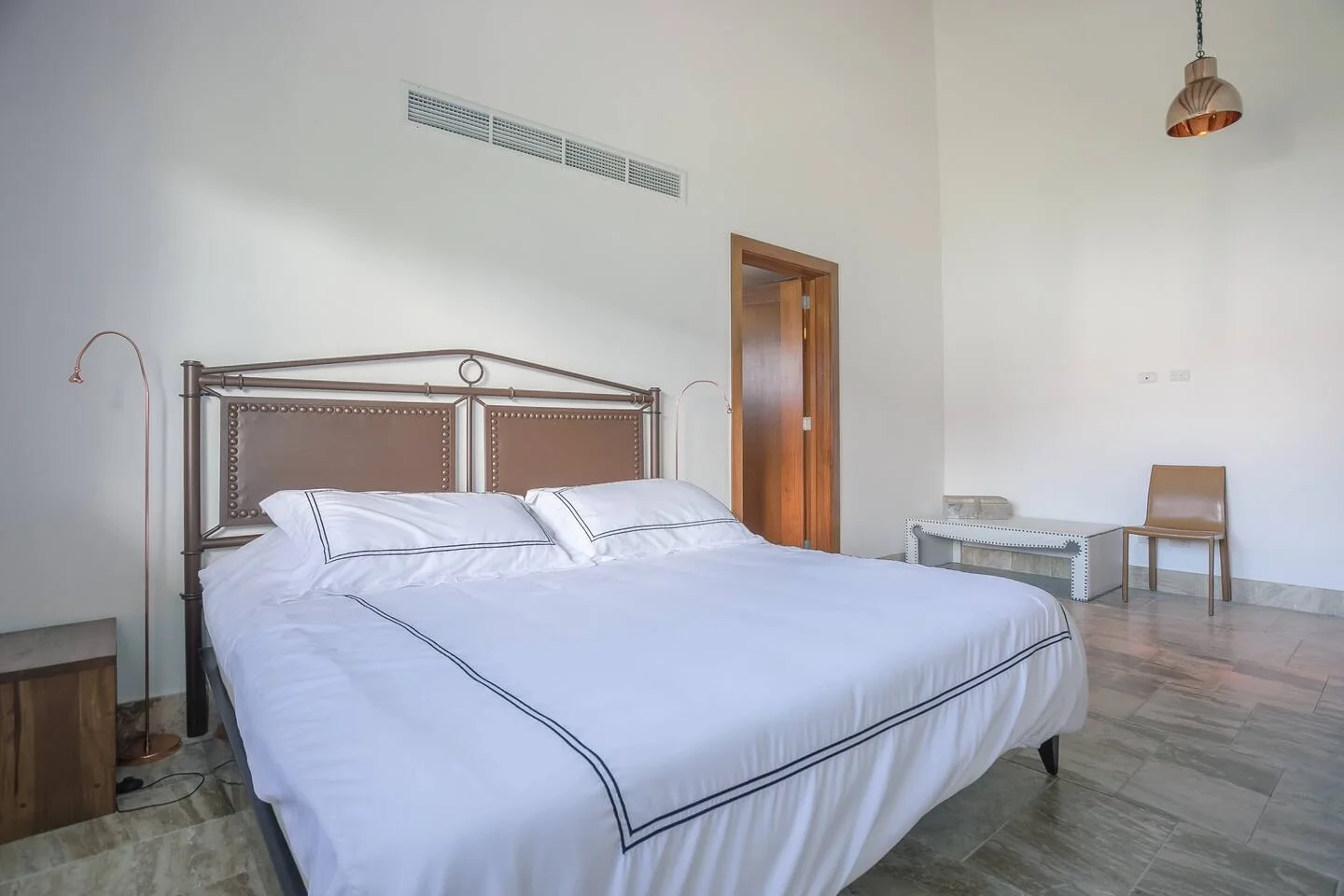 Habitación con cama grande con cabecera de metal y cojines blancos, mesa de madera, silla beige, lámpara de techo de metal en color cobre y paredes blancas.