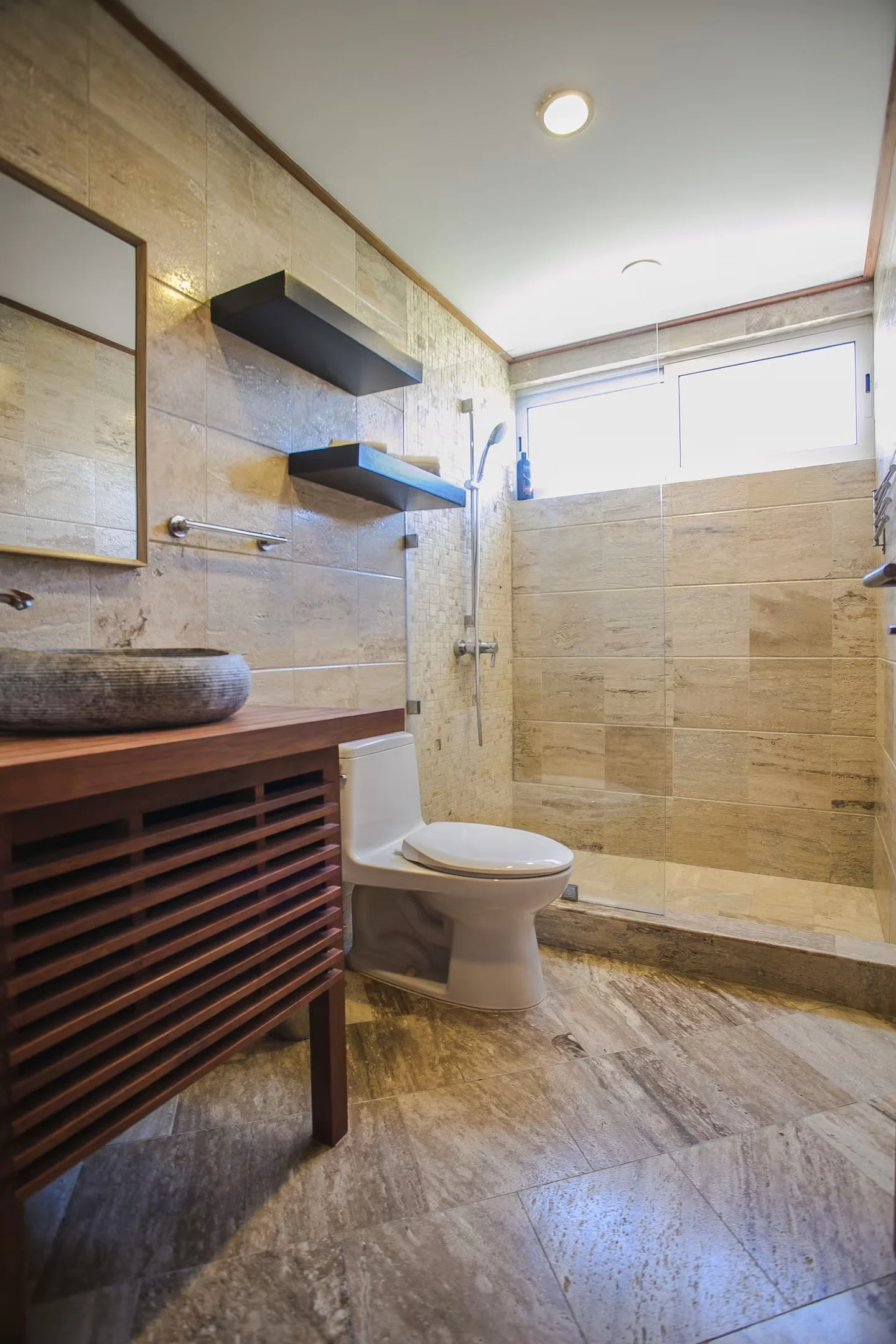 Baño con paredes y piso de piedra, ducha con cristal, inodoro y lavabo de piedra en encimera de madera, con estantes negros en la pared y una ventana grande.