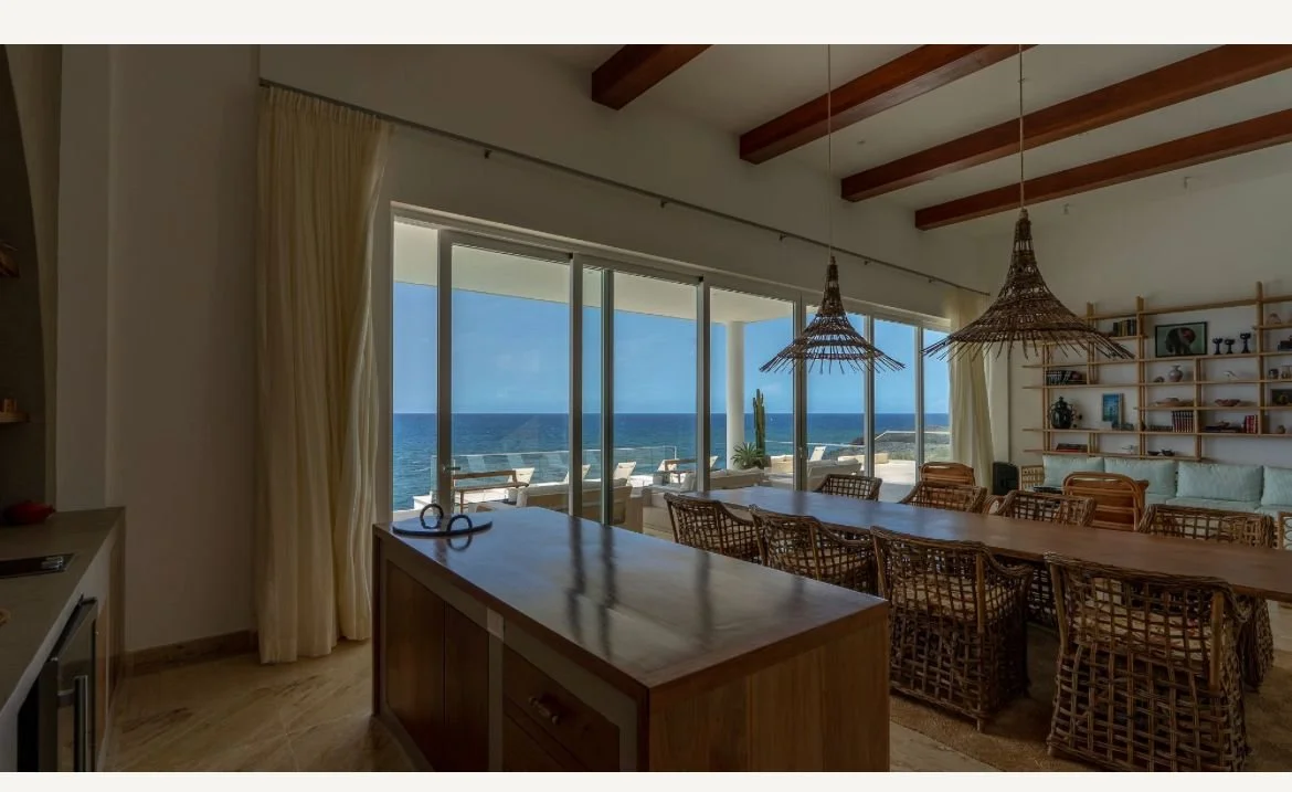 Comedor con mesa de madera y sillas de mimbre, techo con vigas de madera, lámparas colgantes, estanterías con libros, grandes ventanas con vista al mar y una terraza con muebles de exterior.