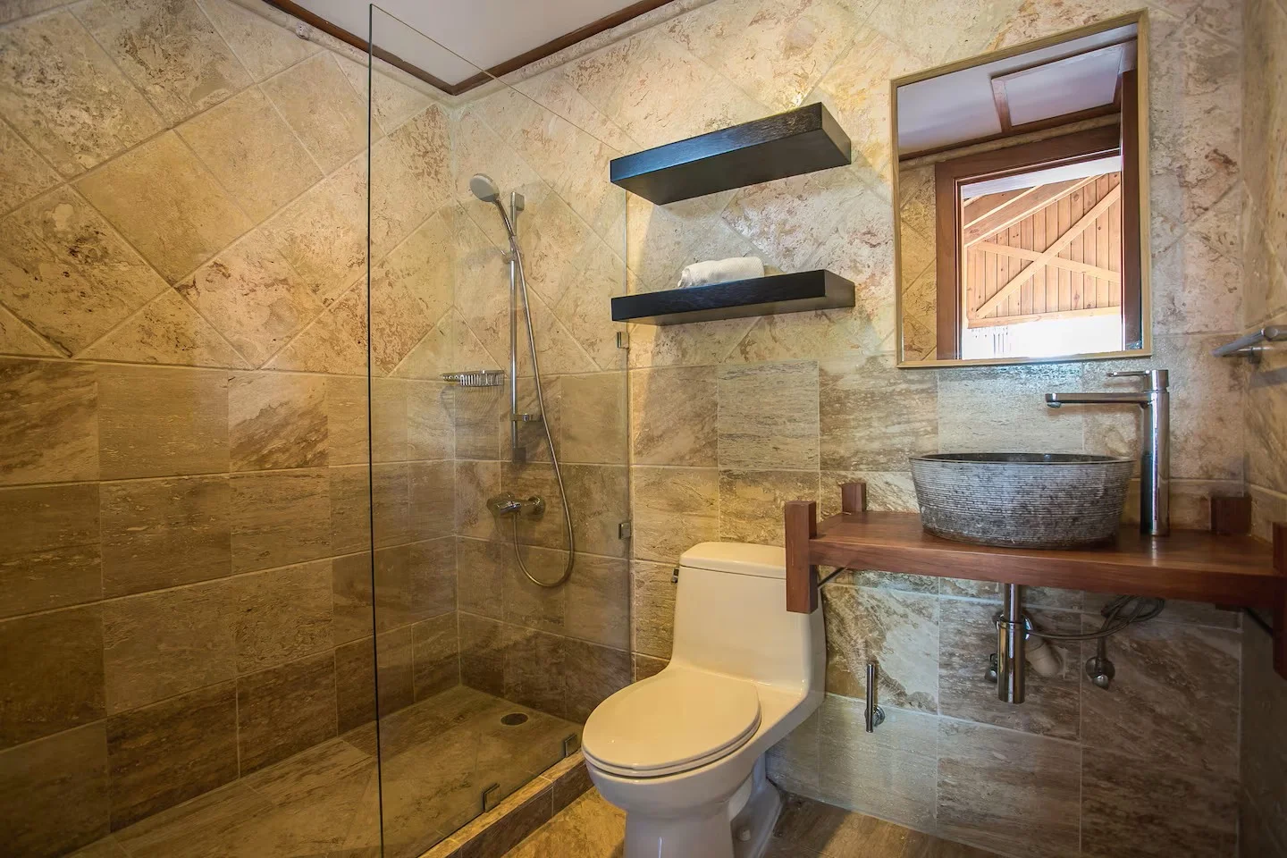 Baño con paredes y piso de piedra en tonos beige, ducha con puerta de vidrio, inodoro blanco, lavabo de piedra en madera, espejo con marco de madera, y ventana que muestra una estructura de madera exterior.