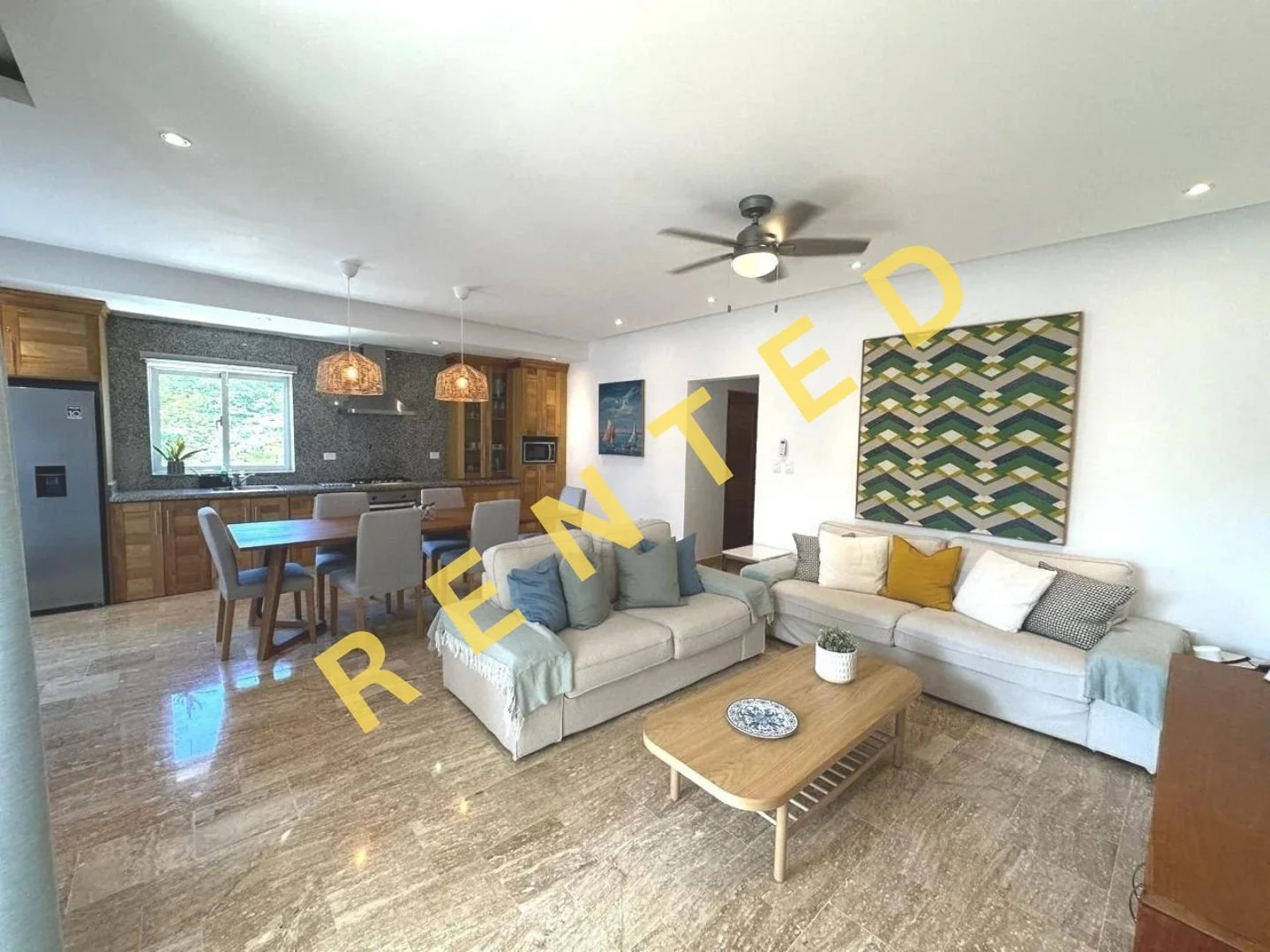 Sala de estar con sofá beige, cojines, mesa de madera y decoración moderna, cocina con comedor en un espacio abierto con ventanas y lámparas colgantes de mimbre.