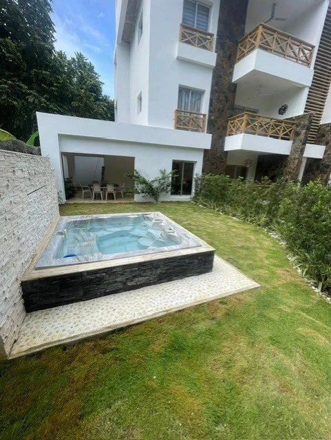 Outdoor hot tub in a garden next to a multi-story building with balconies and a patio area.