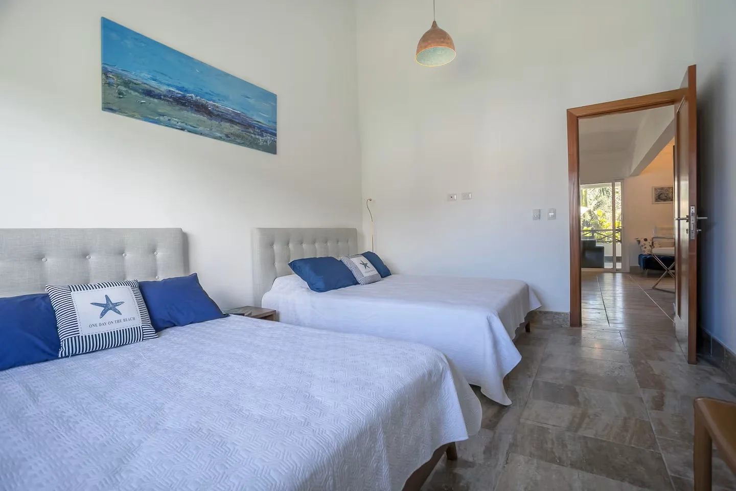 Habitación con dos camas con sábanas blancas y almohadas azules, decorada con tema náutico, tiene una pintura de paisaje marino en la pared y una lámpara colgante de color cobre.