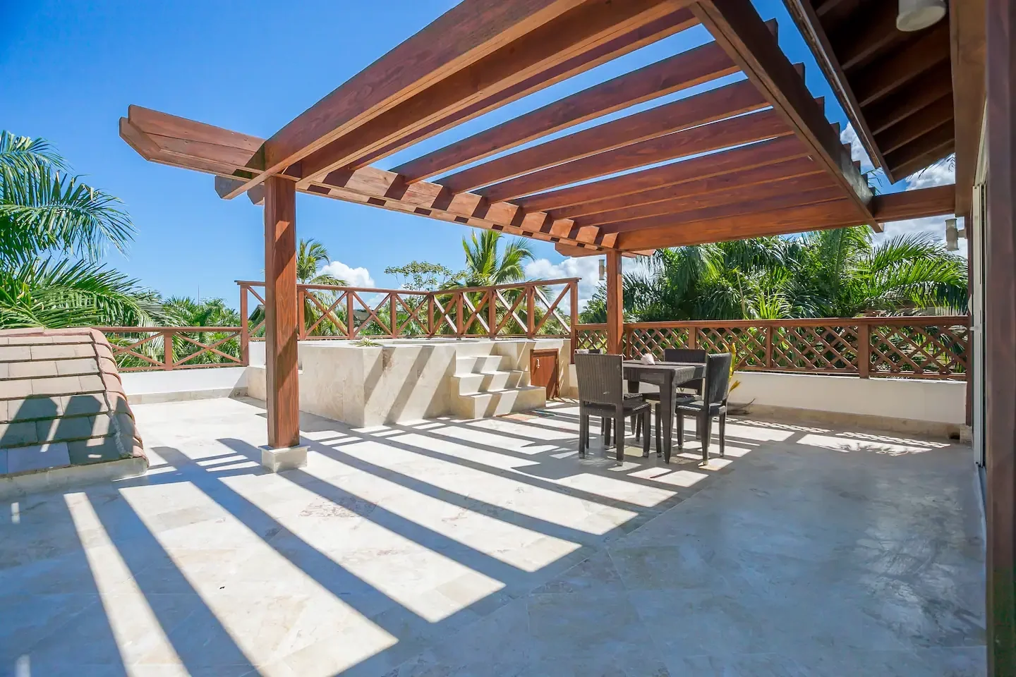 Terraza con sombra de estructura de madera y vista a plantas tropicales bajo cielo azul.