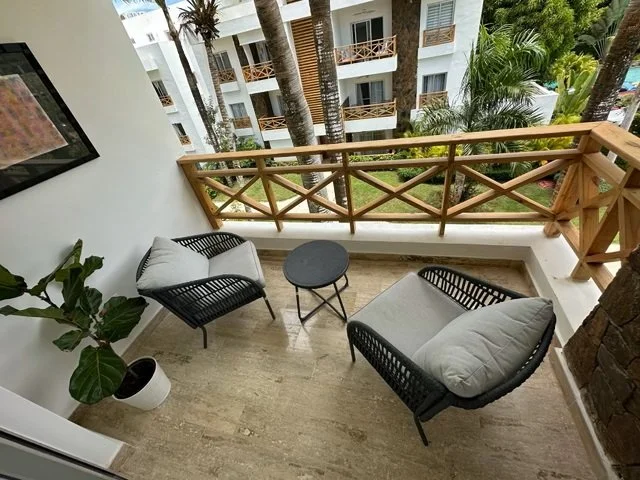 Balcony with two wicker chairs and cushions, a small round table, potted plant, overlooking tropical vegetation and white apartment buildings.