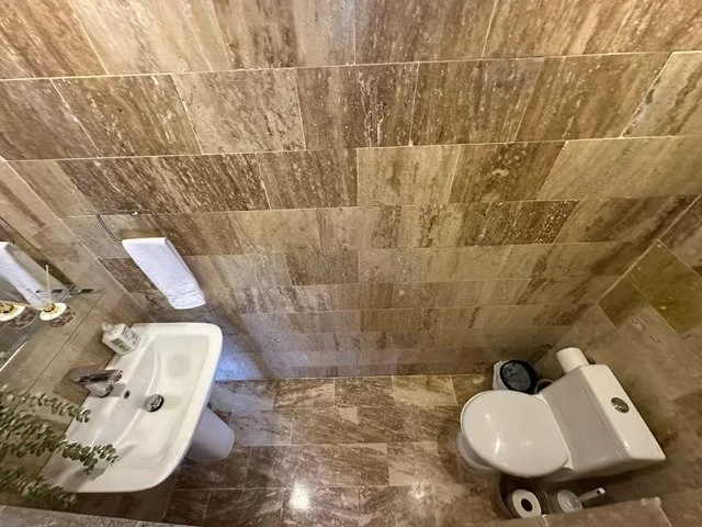 Bathroom with beige marble tiles, featuring a toilet, a white sink, a towel rack with a towel, and a small shelf with toiletries.
