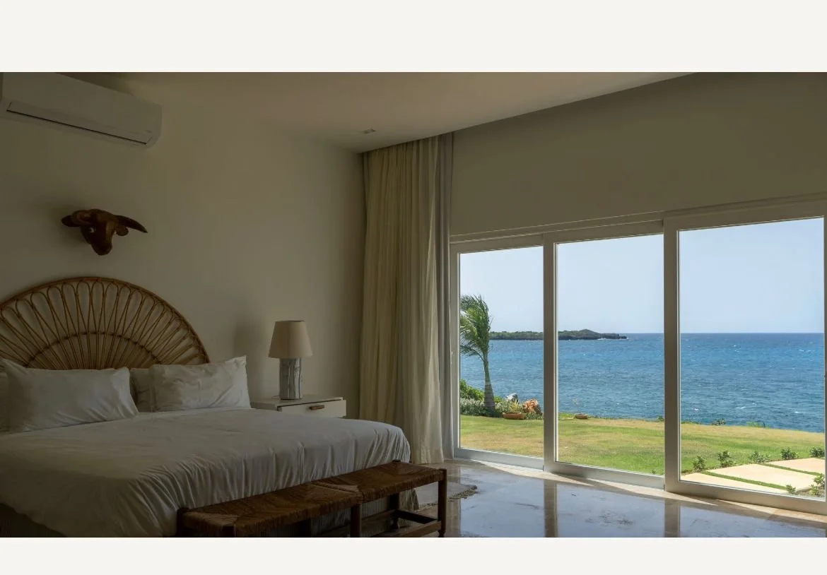 Habitación luminosa con cama grande junto a ventanales que ofrecen vista al mar y jardín. Decoración sencilla con lámpara y elemento decorativo en la pared.