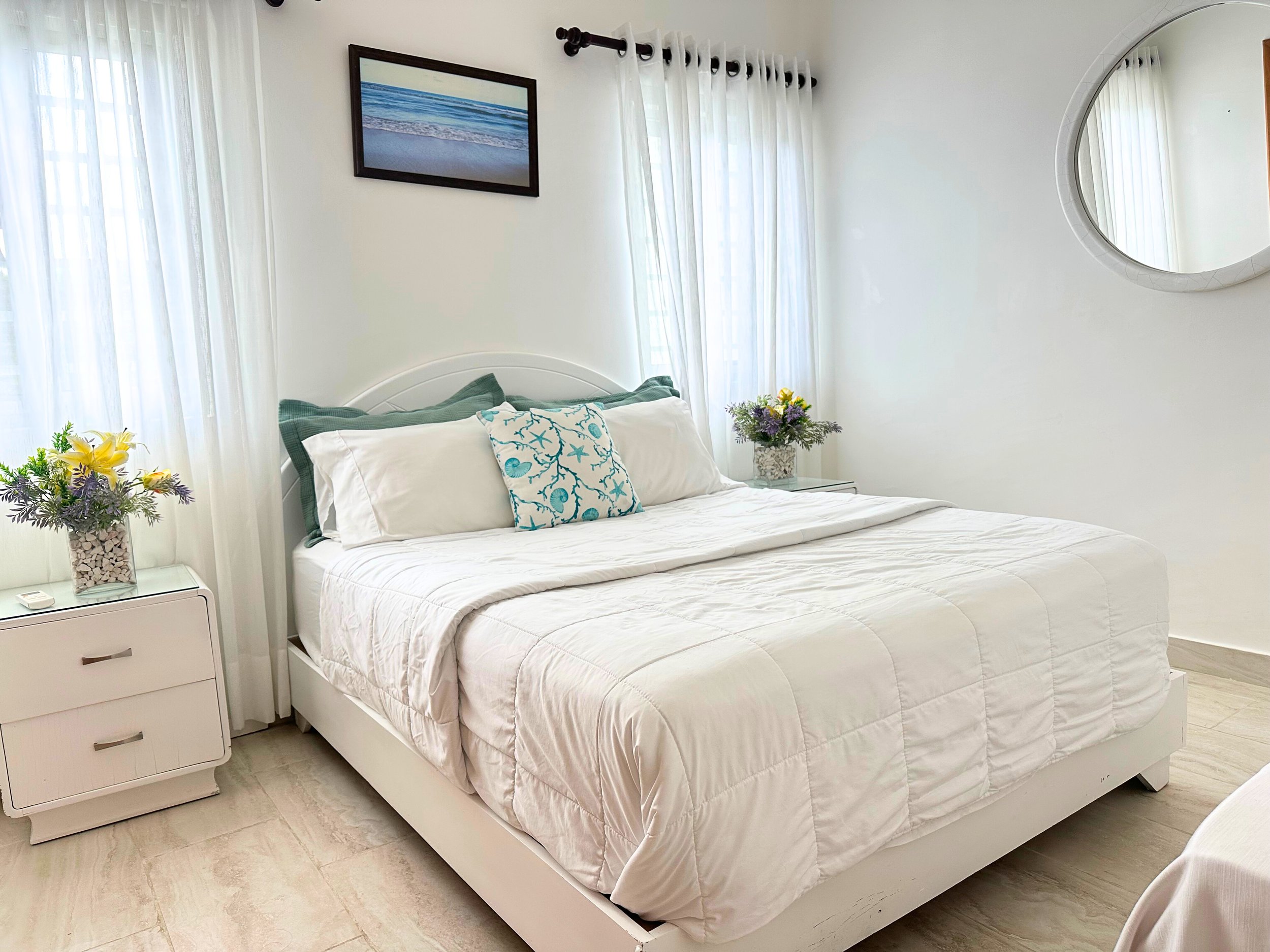 Habitación con cama grande, sábanas blancas, almohadas decorativas, mesas de noche con arreglos florales y cuadro en la pared, cortinas blancas y pared blanca.