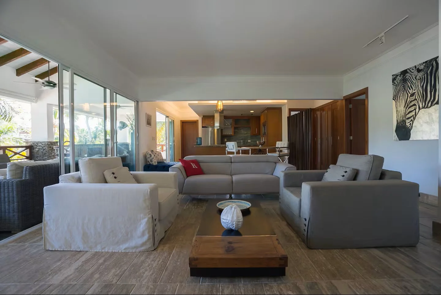 Sala de estar con sillones blancos y grises, mesa de centro de madera, obra de arte de cebras y decoración moderna, con vista a balcón con mobiliario exterior