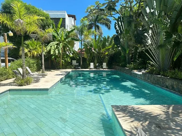 Outdoor swimming pool surrounded by tropical plants and trees, with lounge chairs along the edge on a sunny day.