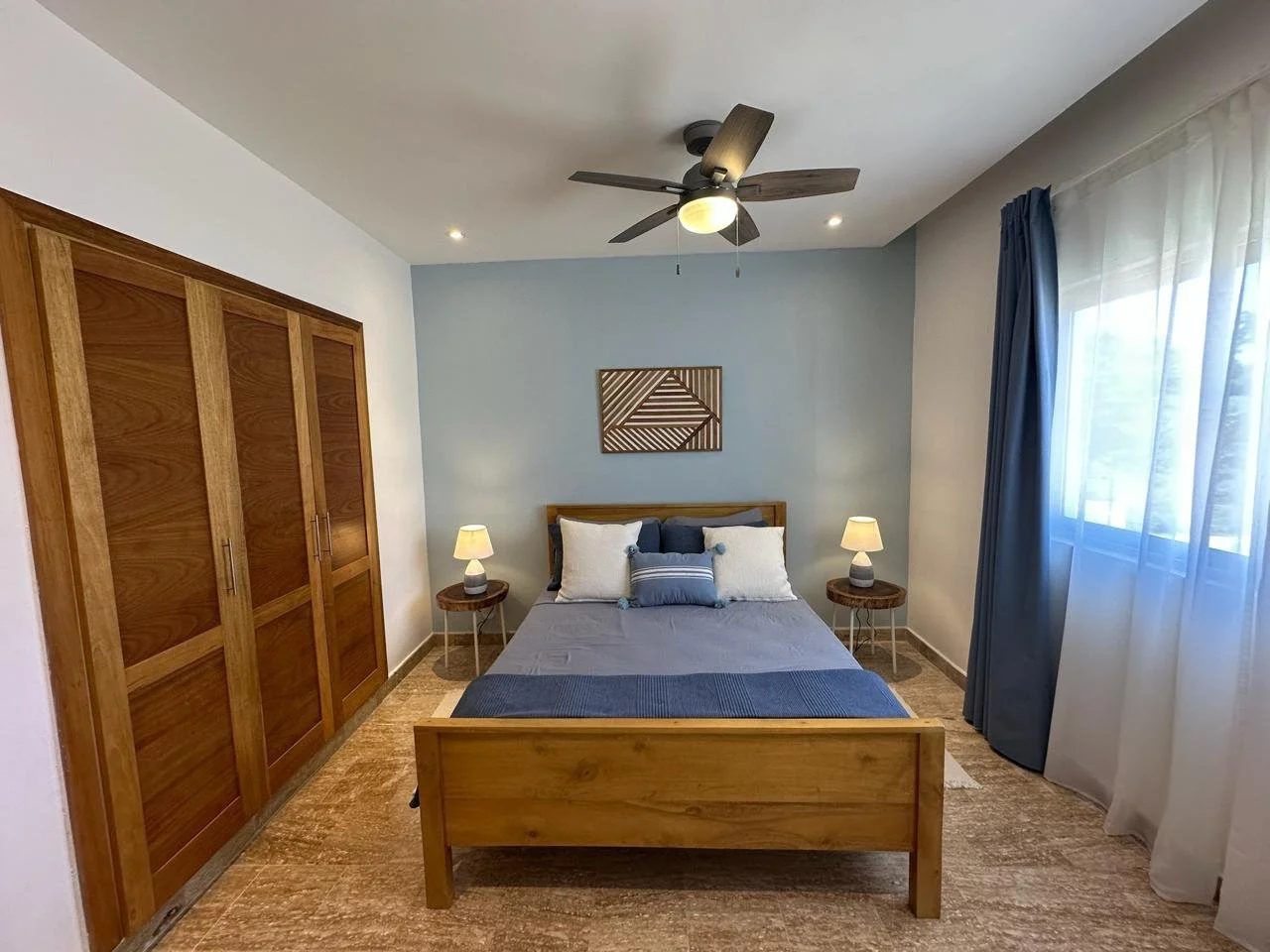 Modern bedroom with wooden bed, blue wall, ceiling fan, large wardrobe, and side tables with lamps. Window with blue curtains on the right.