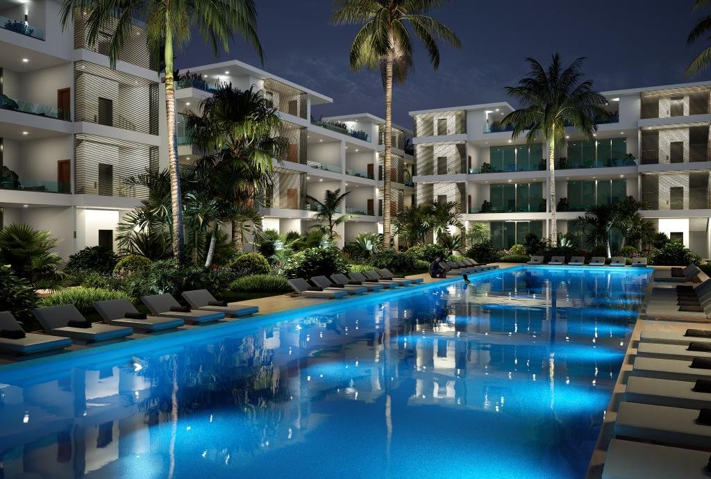 Complejo de apartamentos modernos con piscina iluminada por la noche y palmeras alrededor.
