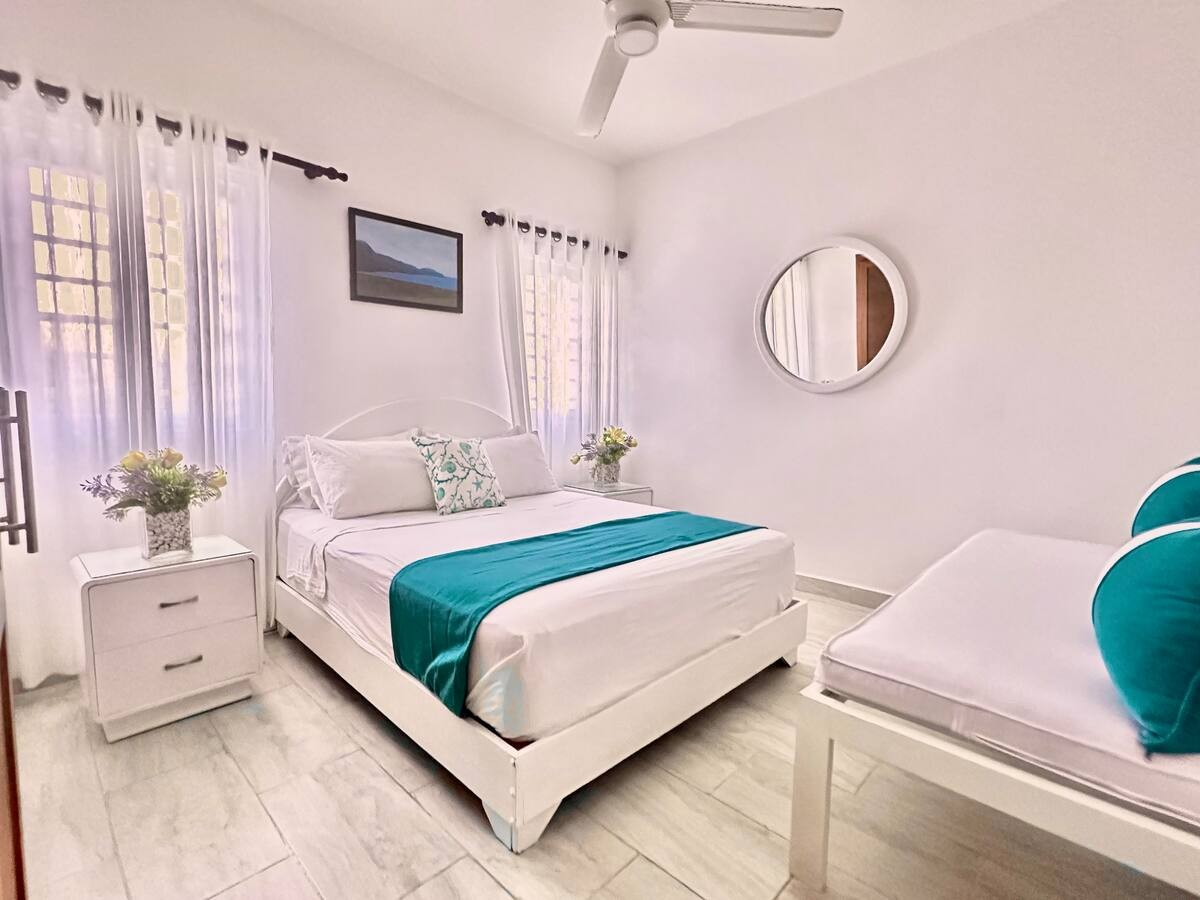 Bright bedroom with a bed featuring white linens and teal accents, a nightstand with flowers, a ceiling fan, mirror, and artwork on the wall.