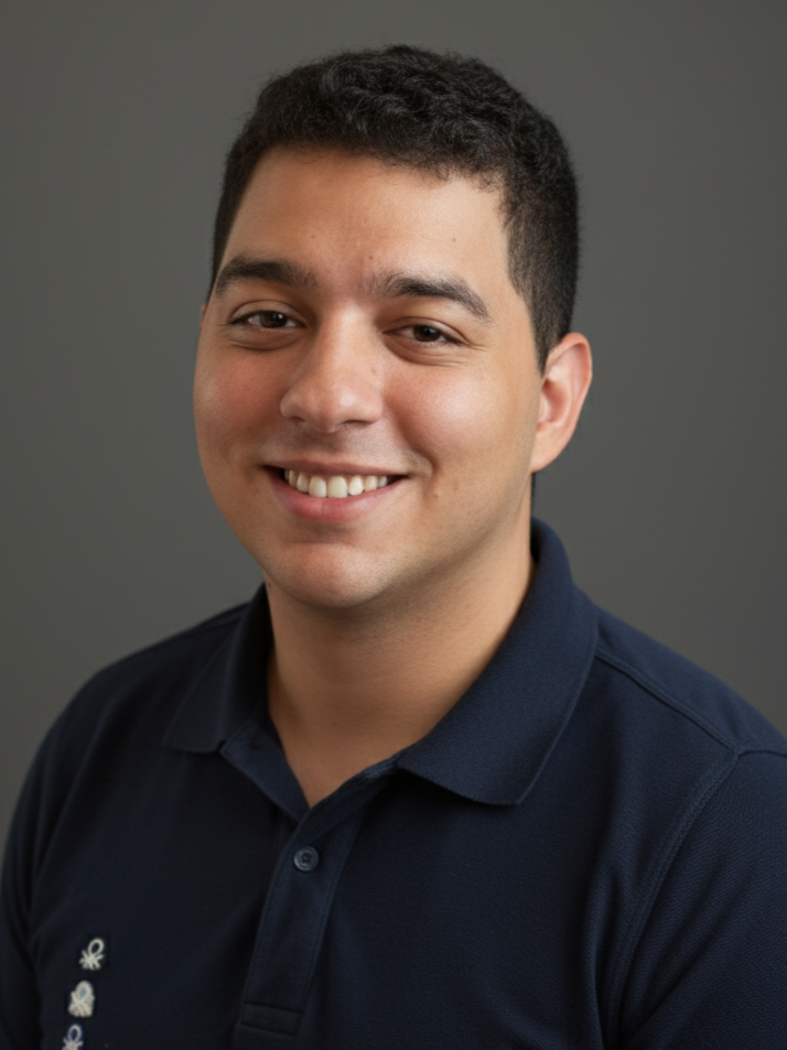 Retrato de un hombre joven sonriendo, viste una camiseta polo de color azul oscuro, con fondo gris.