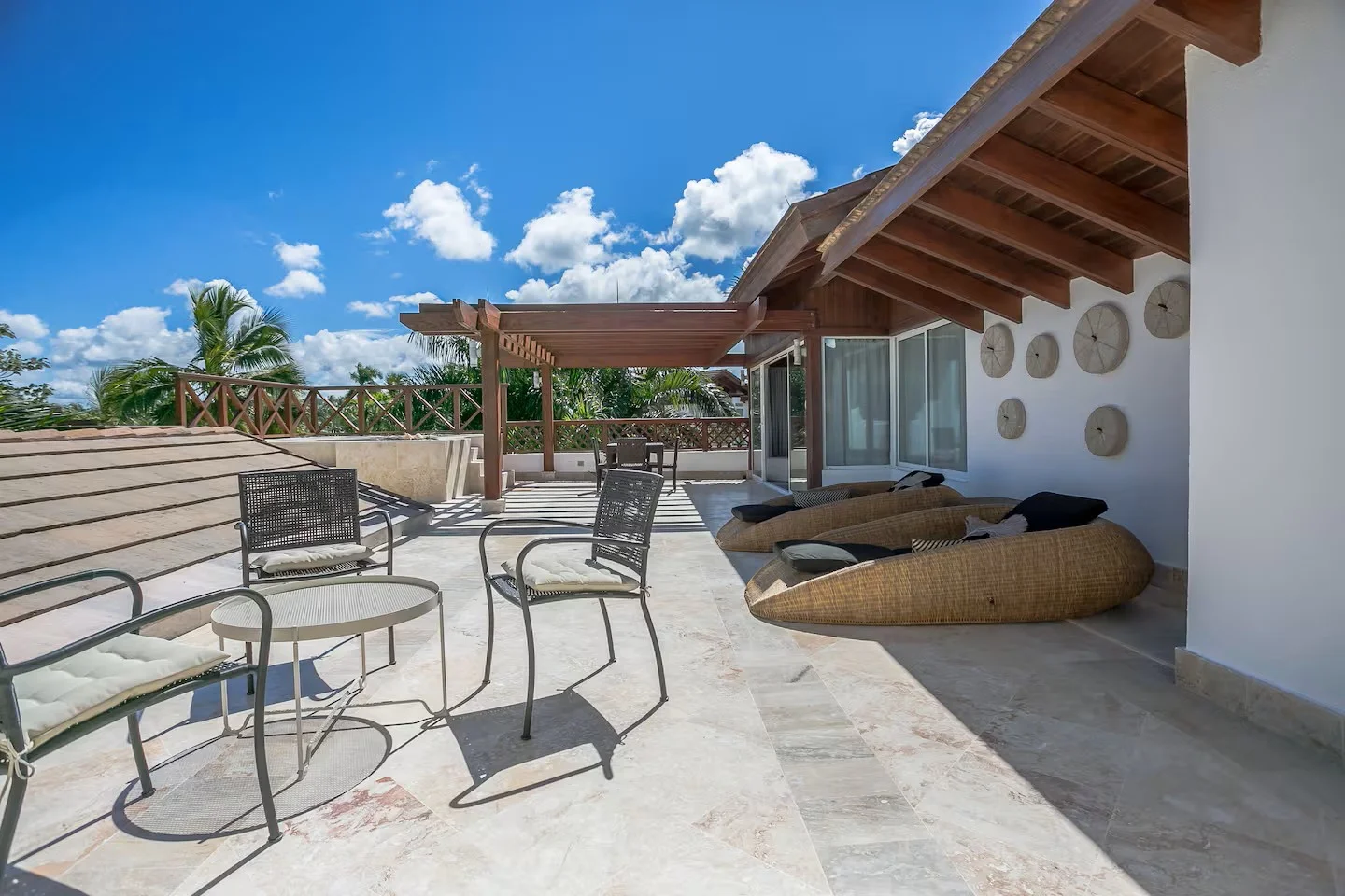 Terraza con sillones y sillas, techo de madera, paredes blancas, decoración de discos en la pared, cielo azul con nubes.