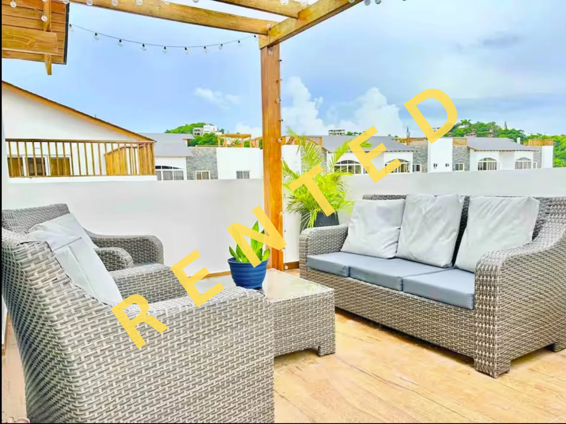 Rincón de terraza con mobiliario de mimbre gris y cojines blancos, planta en maceta azul, paredes blancas, y vista de casas modernas con techos planos en el fondo bajo un cielo nublado.