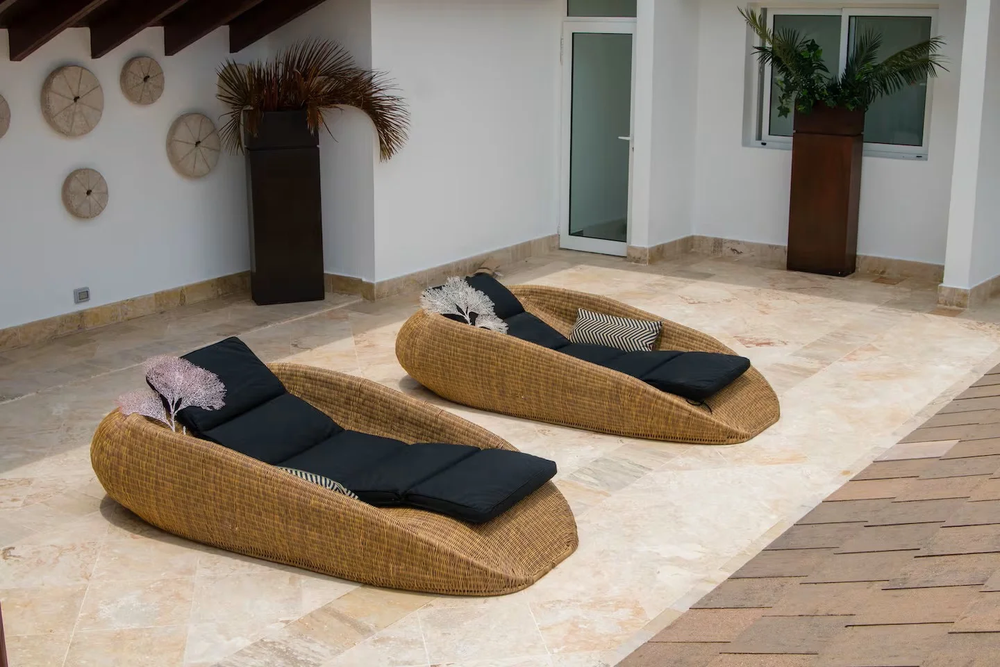 Dos sillas de descanso de mimbre con cojines negros y cojines decorativos, en una terraza con piso de piedra, decorada con plantas en macetas y adornos en la pared.