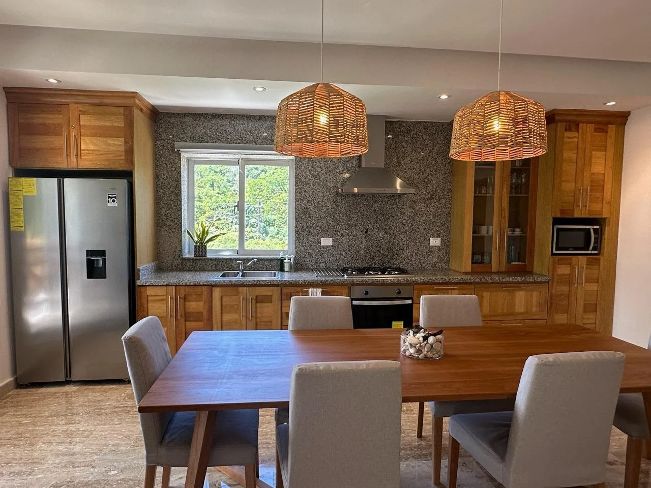 Modern kitchen with wooden cabinets, stainless steel refrigerator, gas stove, and stone countertop. Features a wooden dining table with upholstered chairs and pendant lights above. A window provides natural light, with a small plant on the windowsill