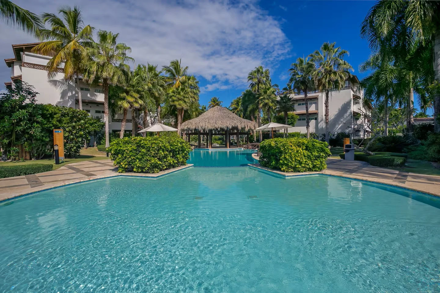 Piscina en un resort con cabaña de palapa y sombrillas, rodeada de palmeras y plantas verdes.