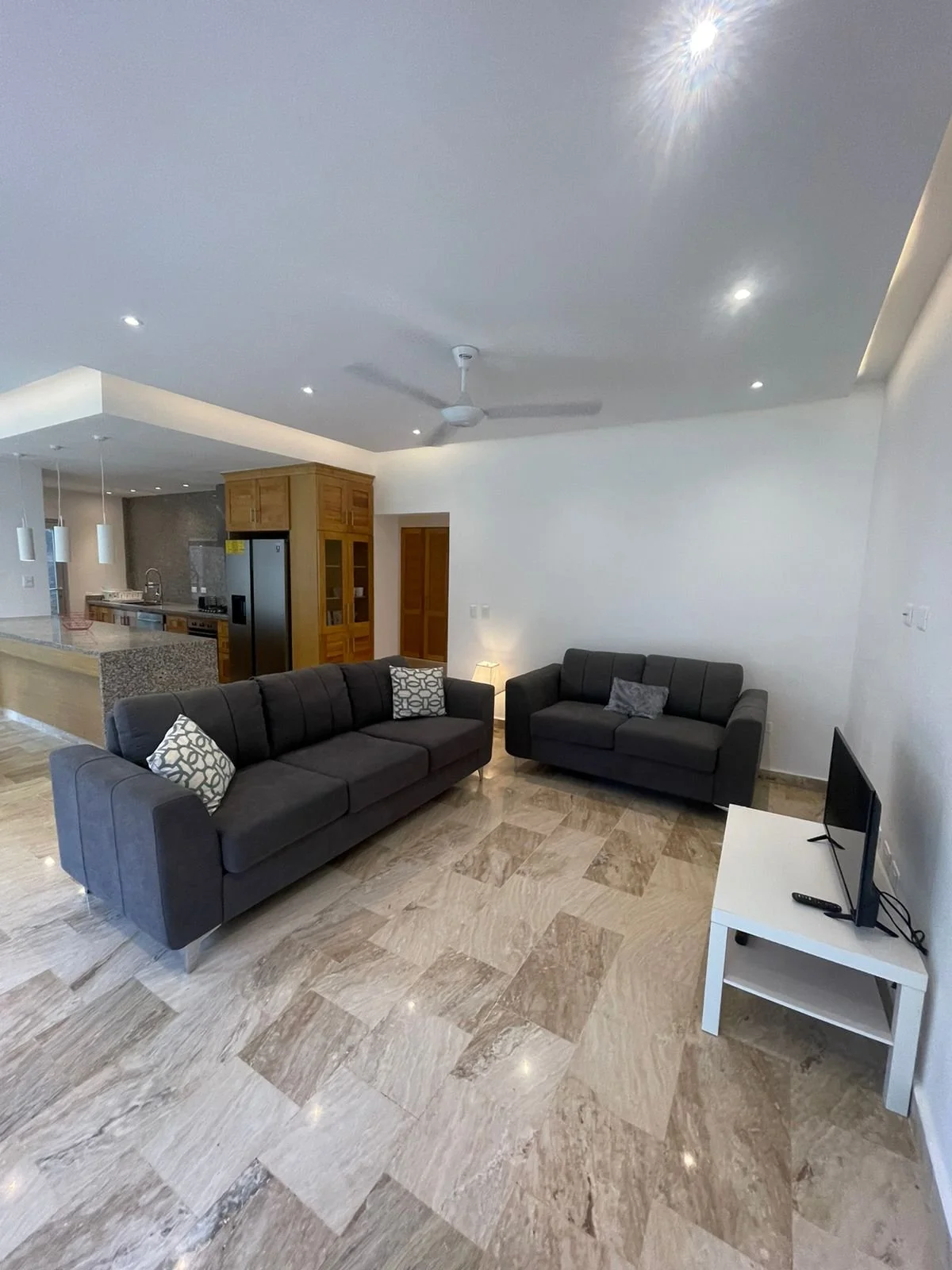 Modern living room with two gray sofas, a small TV on a white stand, beige tiled floor, recessed lighting, ceiling fan, and an open kitchen with wooden cabinets in the background.