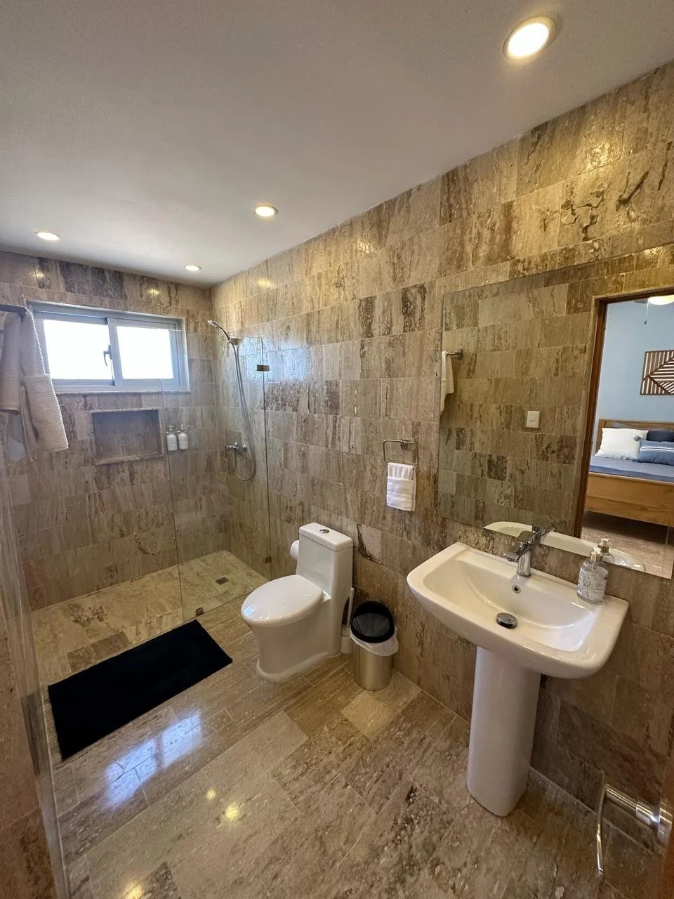 Modern bathroom with beige stone tiles, featuring a walk-in shower with glass partition, a white toilet, a pedestal sink, a mirror, and a small trash can.