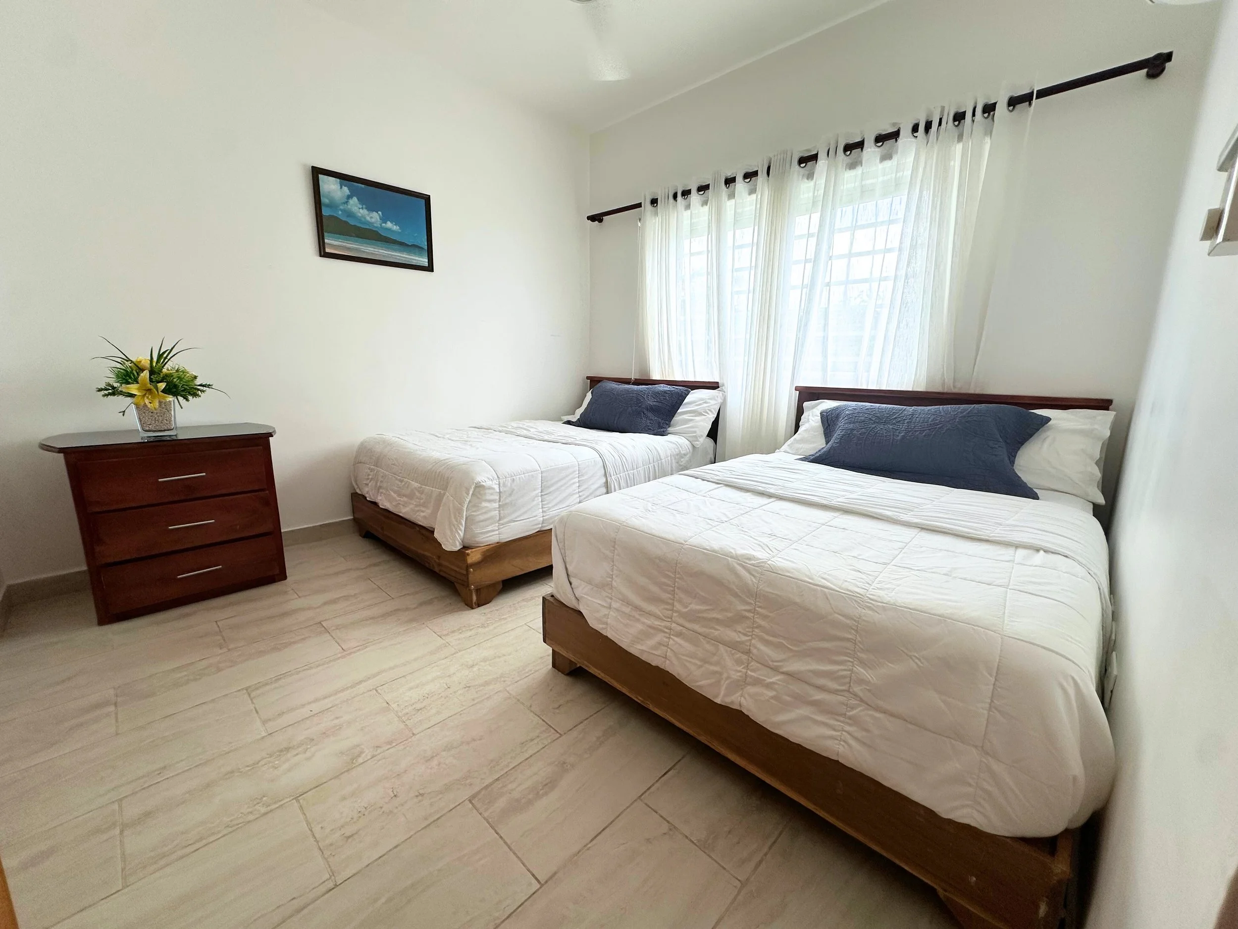 Habitación con dos camas individuales con colchas blancas y almohadas azules, una mesita de noche con florero y flores amarillas, y cortinas blancas en la ventana.