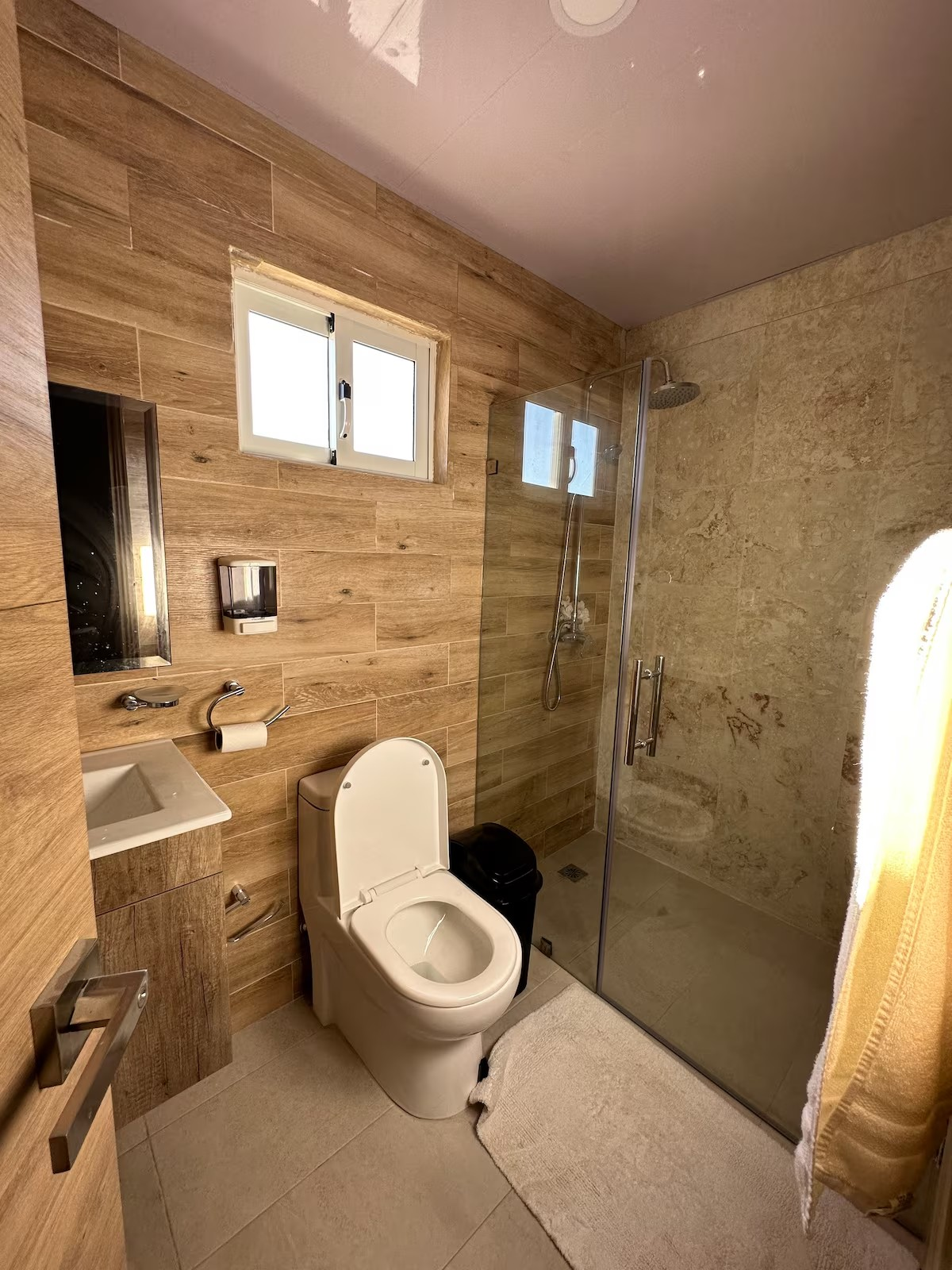 Modern bathroom with a wooden accent wall, toilet, sink, trash can, and a glass shower enclosure.