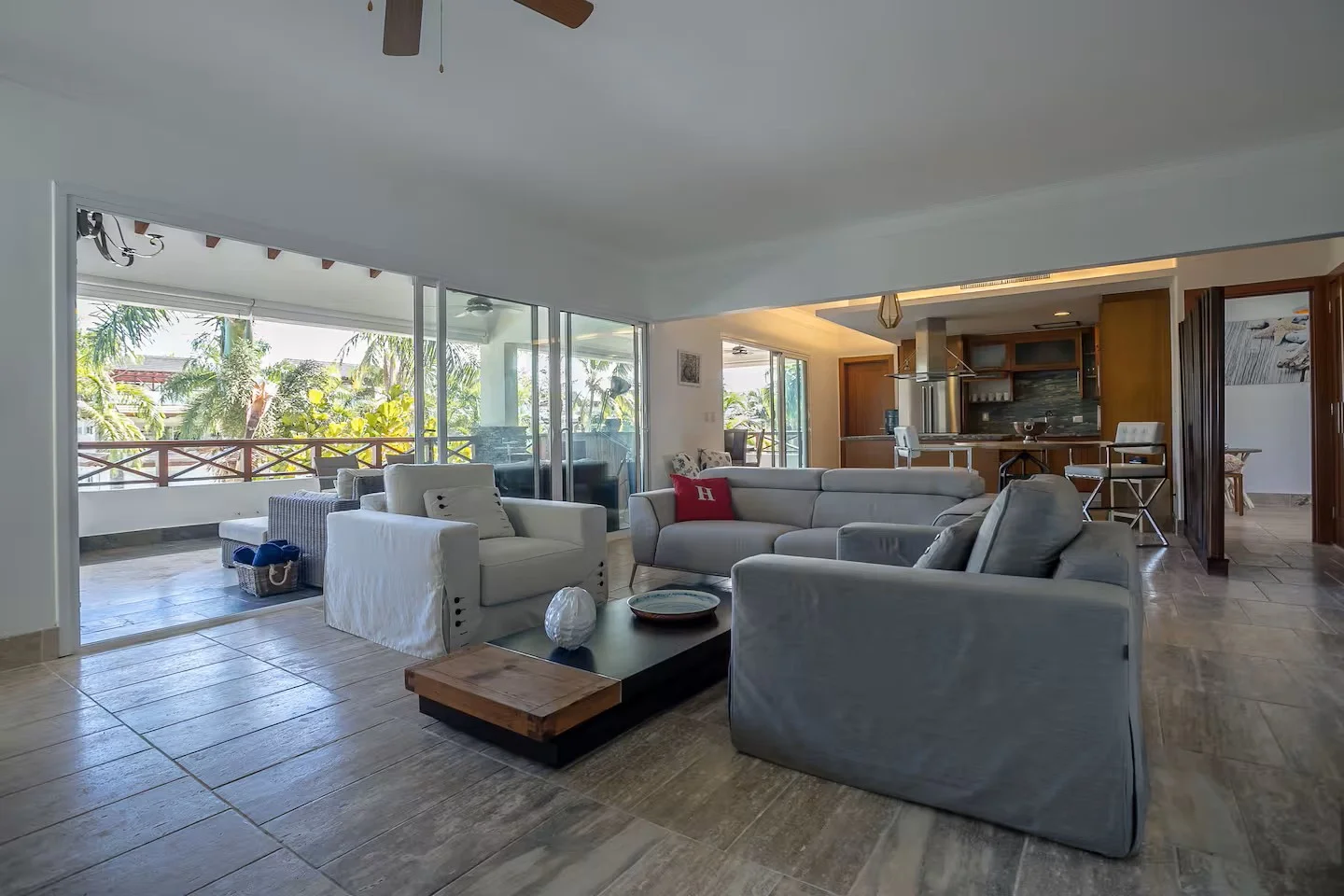 Sala de estar con sillones grises, mesas de centro, grandes ventanas con vista a un balcón con plantas tropicales, cocina en el fondo, decoración moderna.