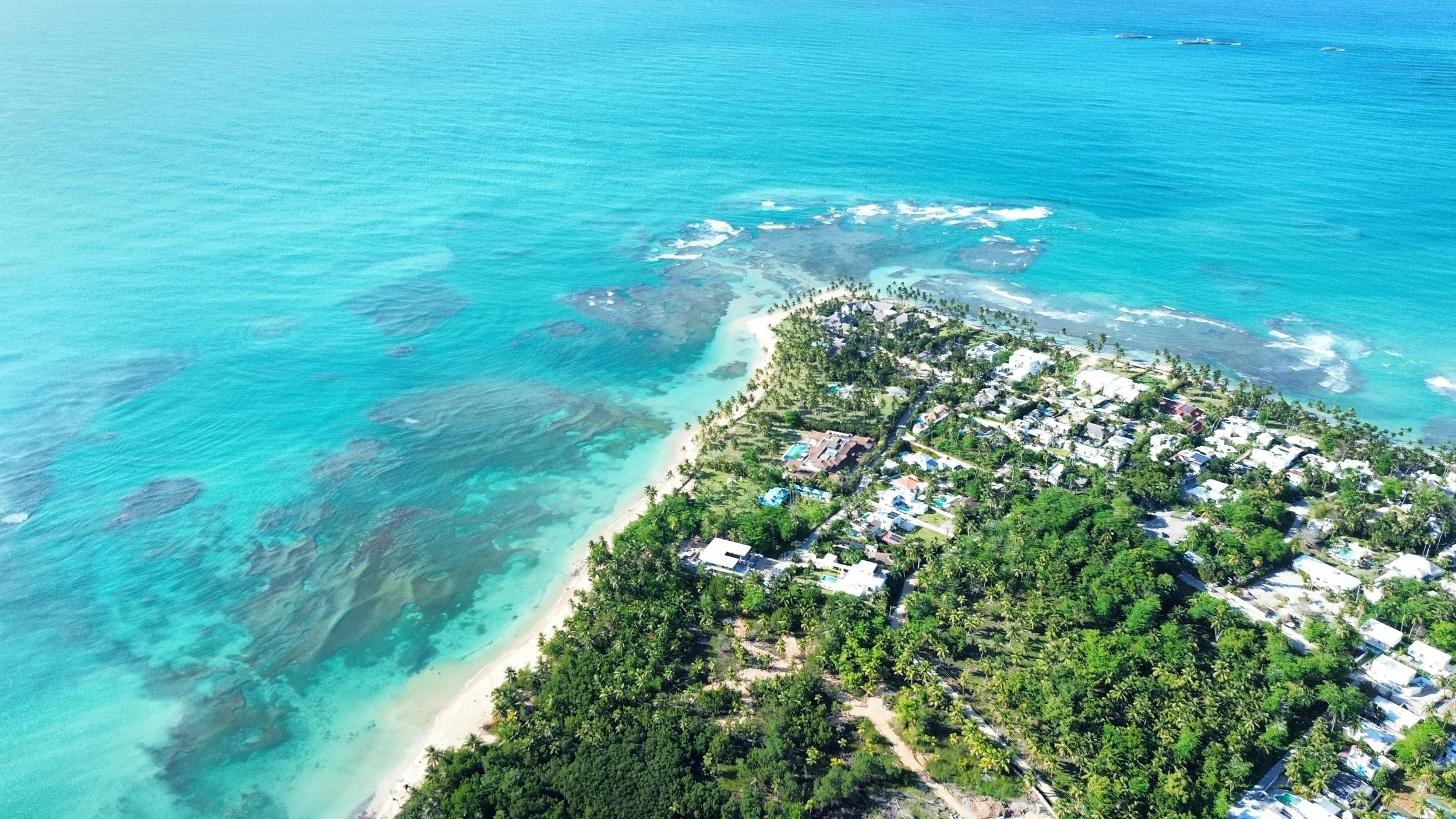 Vista aérea de una costa con aguas turquesas, playa de arena clara y una comunidad rodeada de vegetación y palmeras.