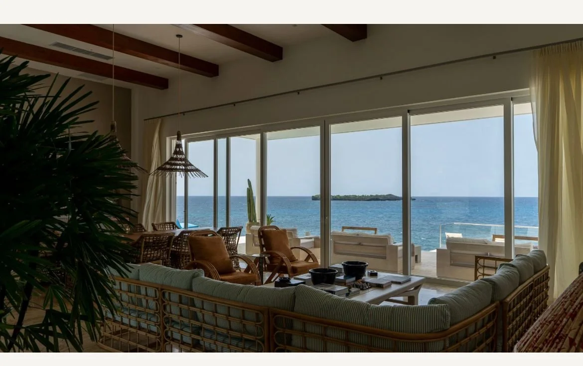 Sala de estar con muebles de mimbre, vista al océano a través de grandes ventanales y techo con vigas de madera.