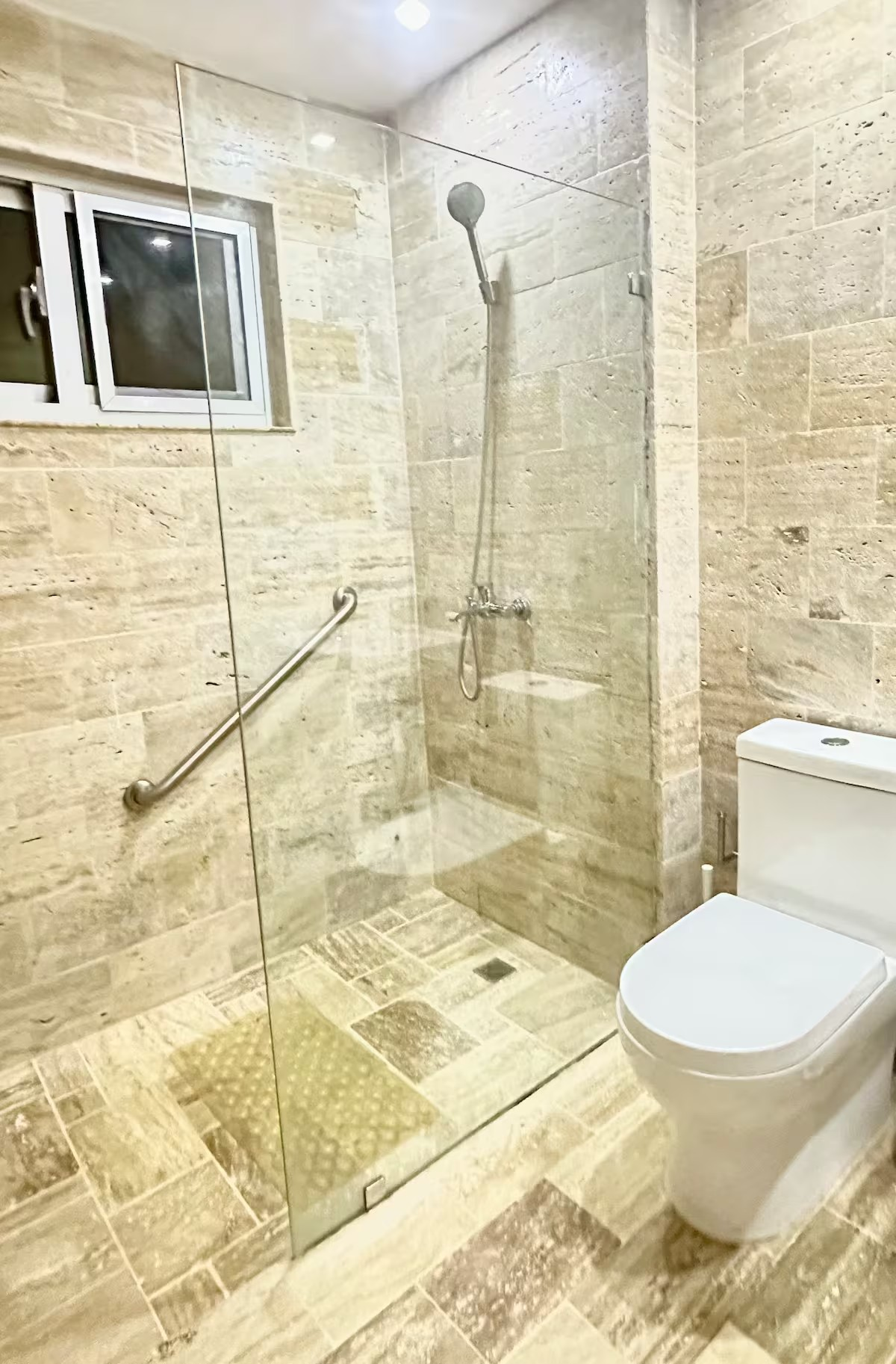 Modern bathroom with a glass shower enclosure, handheld showerhead, beige stone tiles, small window, wall-mounted grab bar, and a toilet.