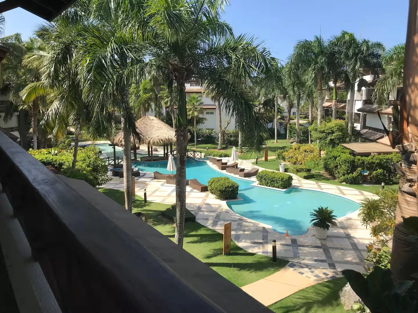 Área de piscina en un resort, rodeada de palmeras, hamacas, sombrillas, un pabellón de bambú, y jardines verdes.