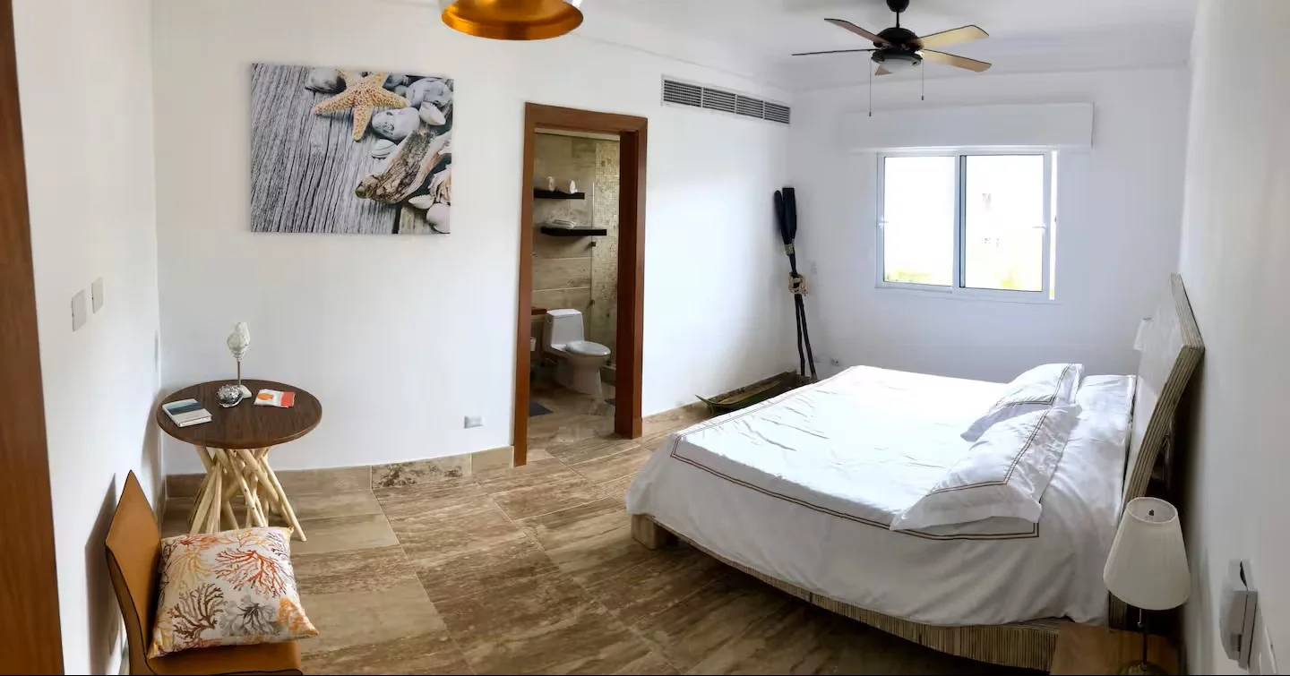 Habitación con cama, ventana, lámpara, mural de conchas, baño en puerta opuesta, piso de piedra y ventilador en el techo.