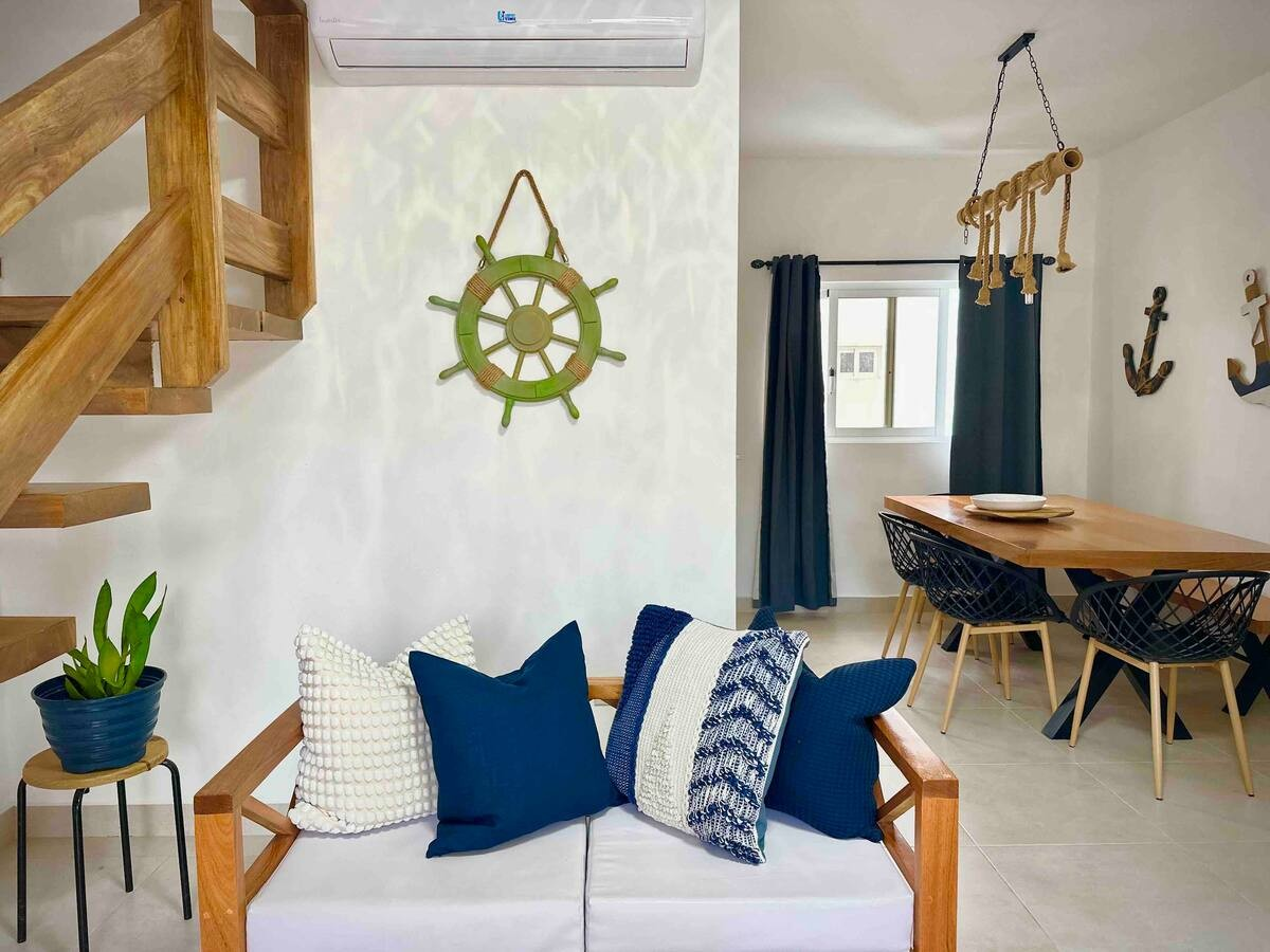 Modern living and dining room with nautical decor, featuring a wooden staircase, blue and white couch with pillows, a wall-mounted ship wheel, rope lighting fixture, dining table, and chairs.
