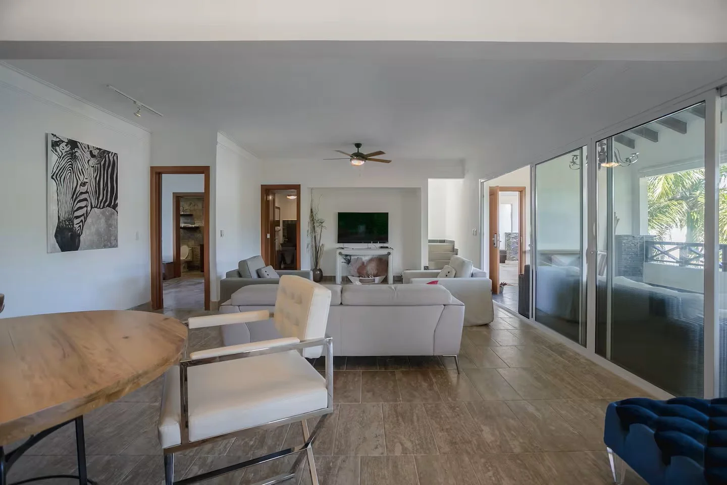 Sala de estar moderna con paredes blancas, pisos de madera y un gran ventanal de vidrio. Muebles blancos, una televisión en la pared y decoración con plantas y cuadros.