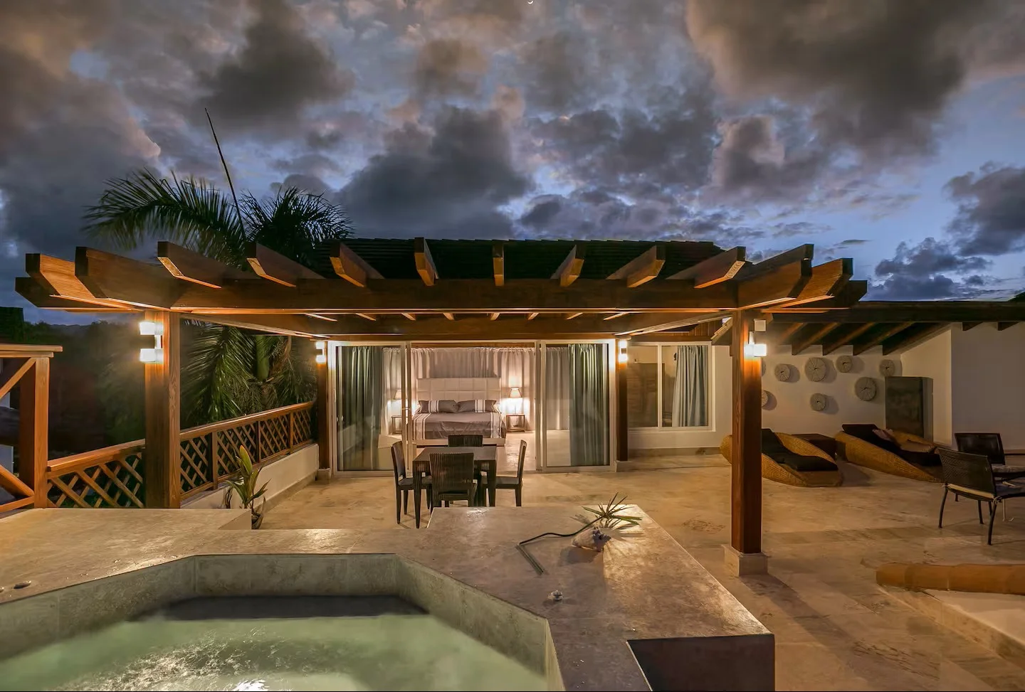 Terraza con jacuzzi y mobiliario, vista a la habitación con cama y cortinas, con noche oscura y cielo con nubes.
