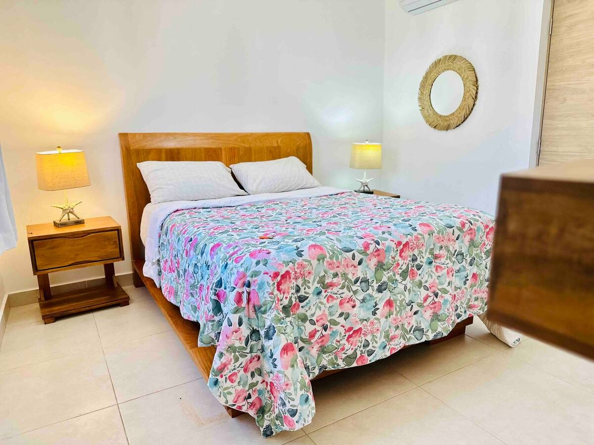 Bedroom with wooden bed frame, floral bedspread, two bedside tables with lamps, and a round decorative mirror on the wall.