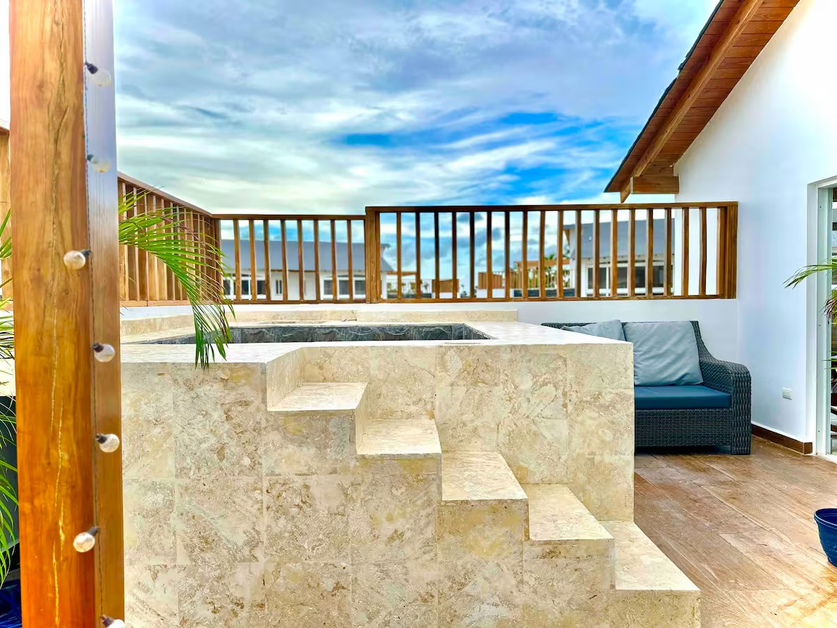 Outdoor rooftop terrace with a small built-in hot tub, stone steps, and wooden railing. There are potted plants and a wicker sofa with cushions. The sky is blue and cloudy.