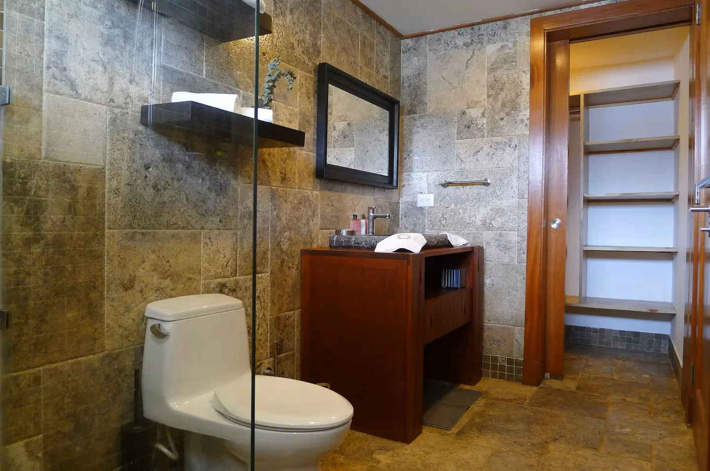 Baño con paredes de piedra y piso de piedra en tonos marrones, lavabo de madera oscura con encimera de piedra, espejo rectangular negro, inodoro blanco, estantería abierta en la puerta, y objetos de tocador en el lavabo.